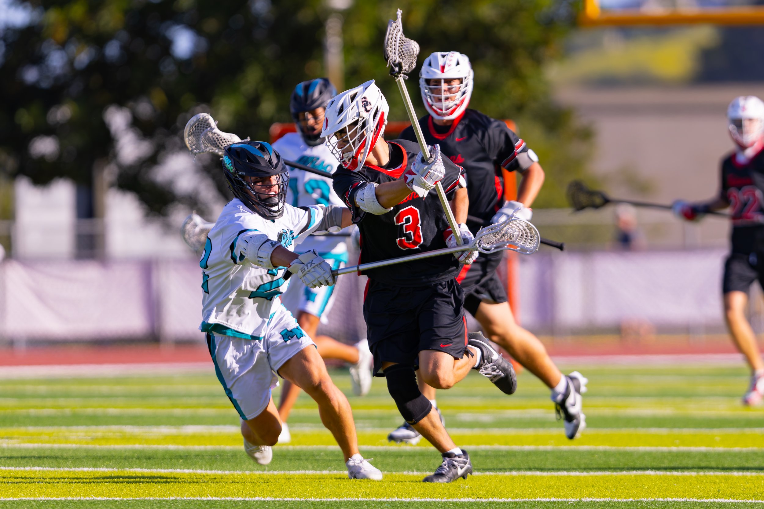 Lacrosse players in action on the field, wearing helmets and uniforms, with one player in black and red tightly holding the ball while others attempt to defend and intercept.