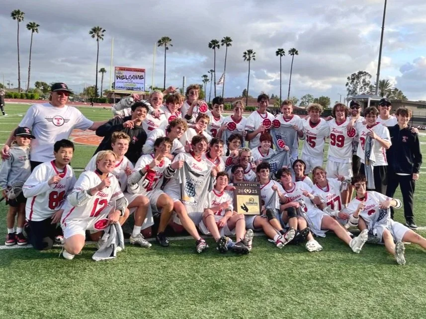 A group of young boys and their coaches on a sports field celebrating after a game, wearing white jerseys, some holding towels and a trophy, with palm trees in the background.