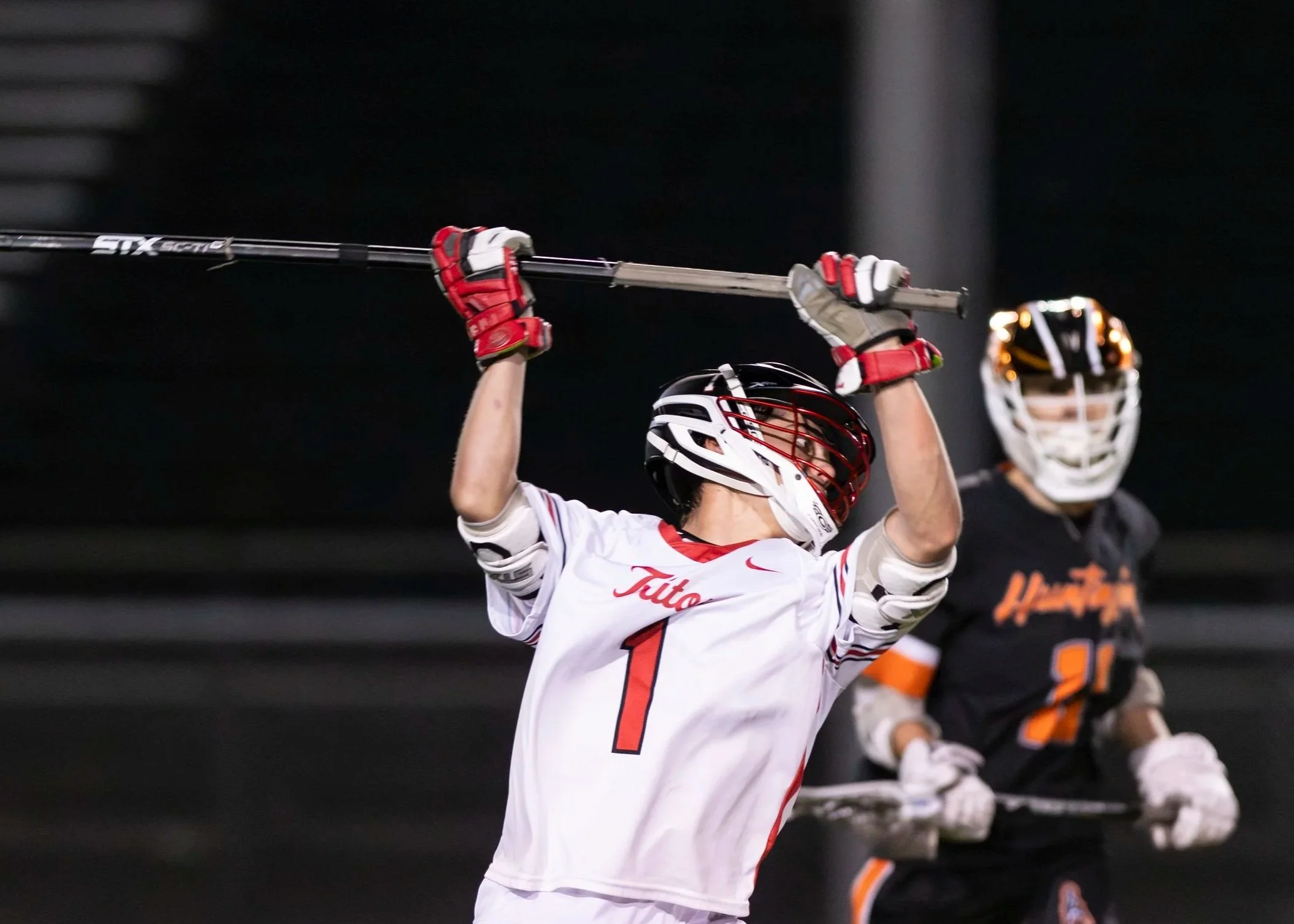 Lacrosse player in white jersey with number 1, holding lacrosse stick overhead during game, with opposing player in dark jersey in background. Nighttime sports setting.