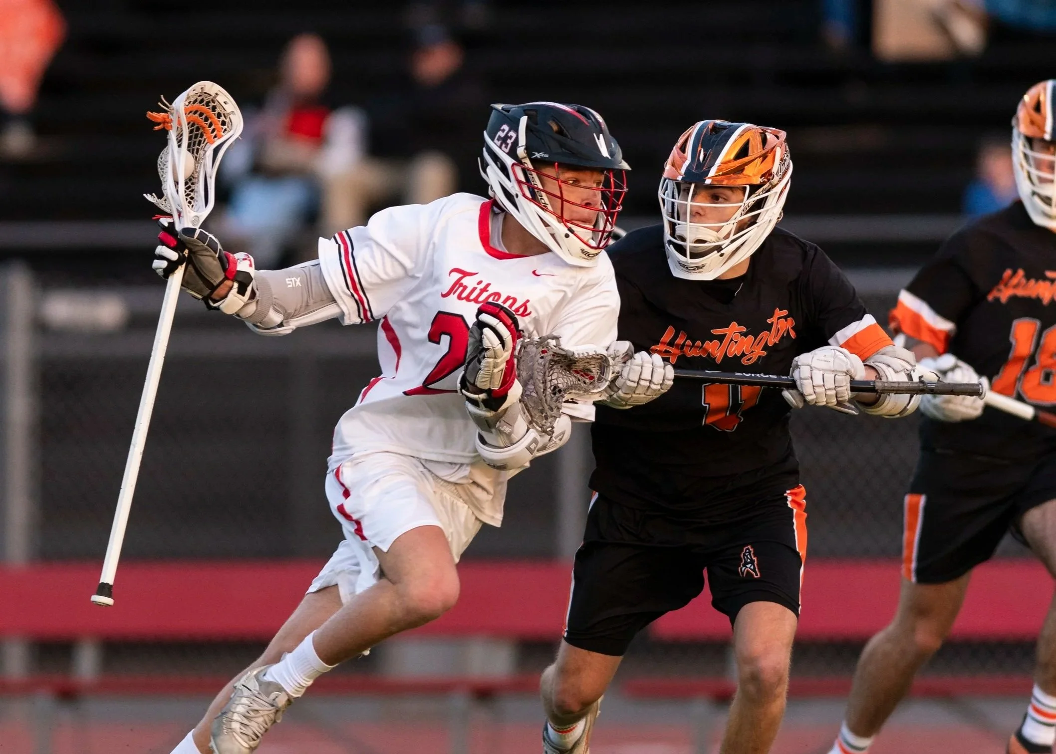 Lacrosse players in action on the field, with one player in a white jersey and two in black jerseys competing for the ball, with a green turf field and spectators in the background.