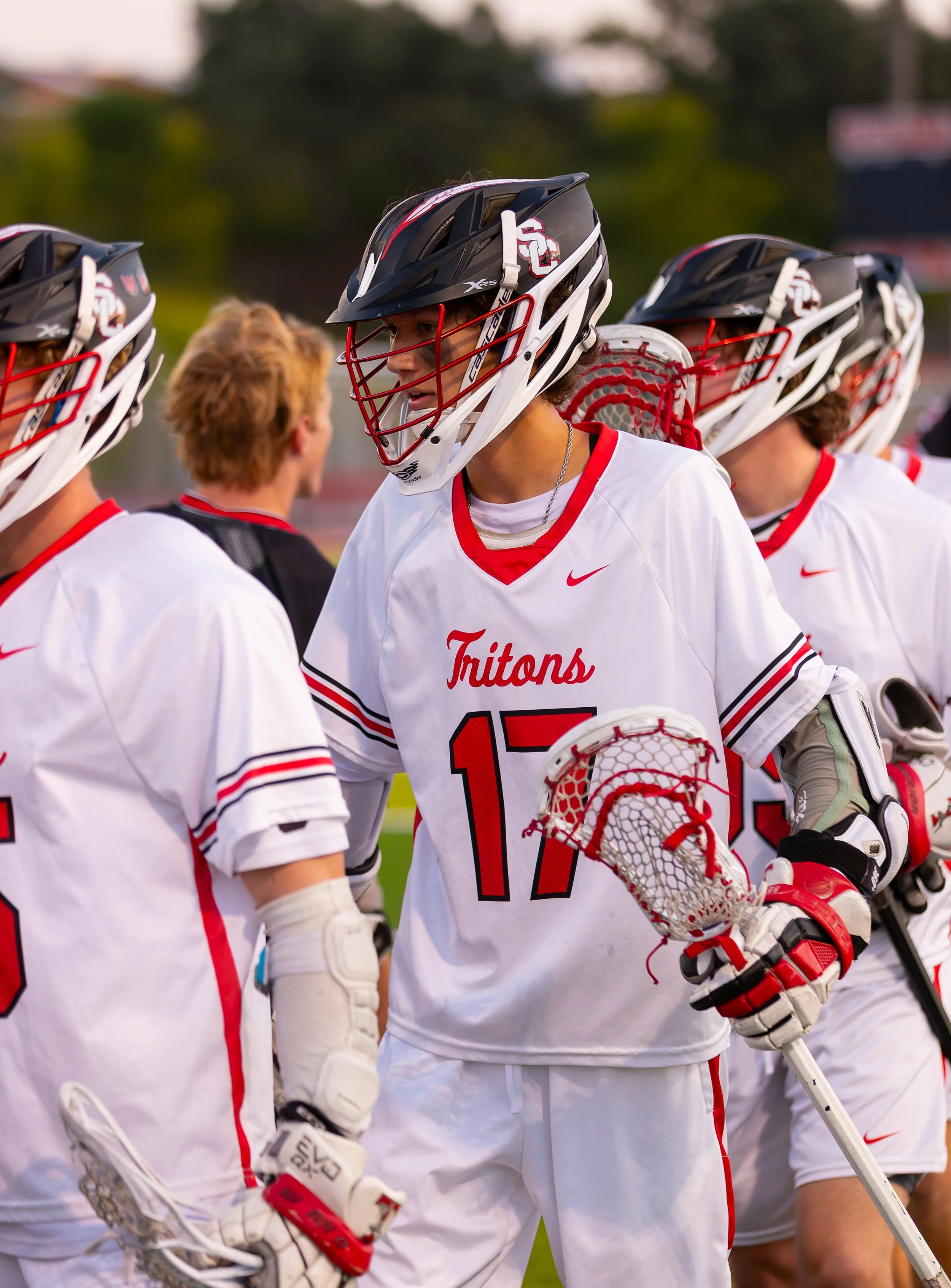 Lacrosse players wearing white jerseys with red accents and helmets gathered on the field, engaging in a discussion during a game.