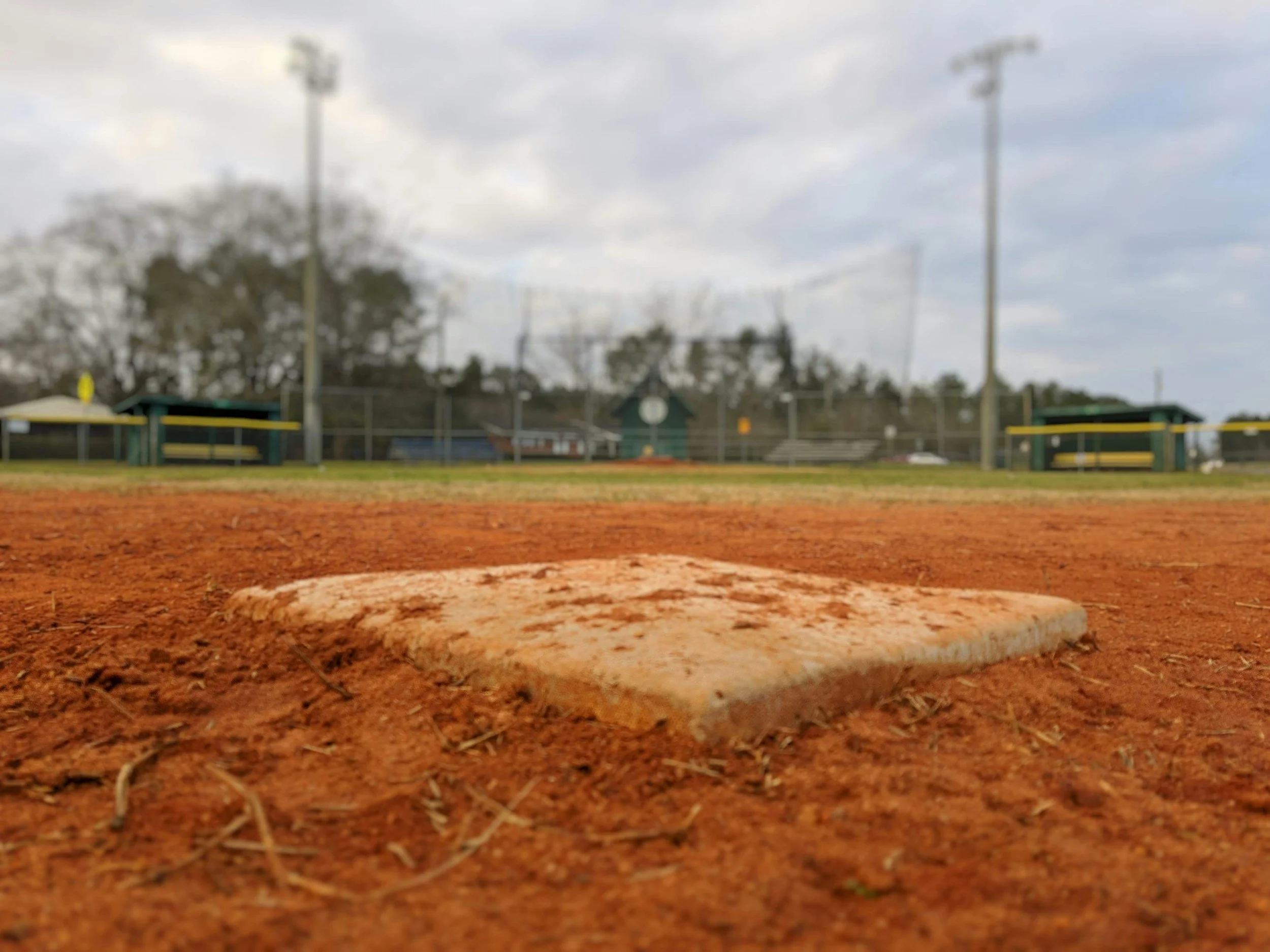 Close-up of a baseball home plate on a dirt field in Lincoln County, Oregon with a baseball field in the background, under a cloudy sky.