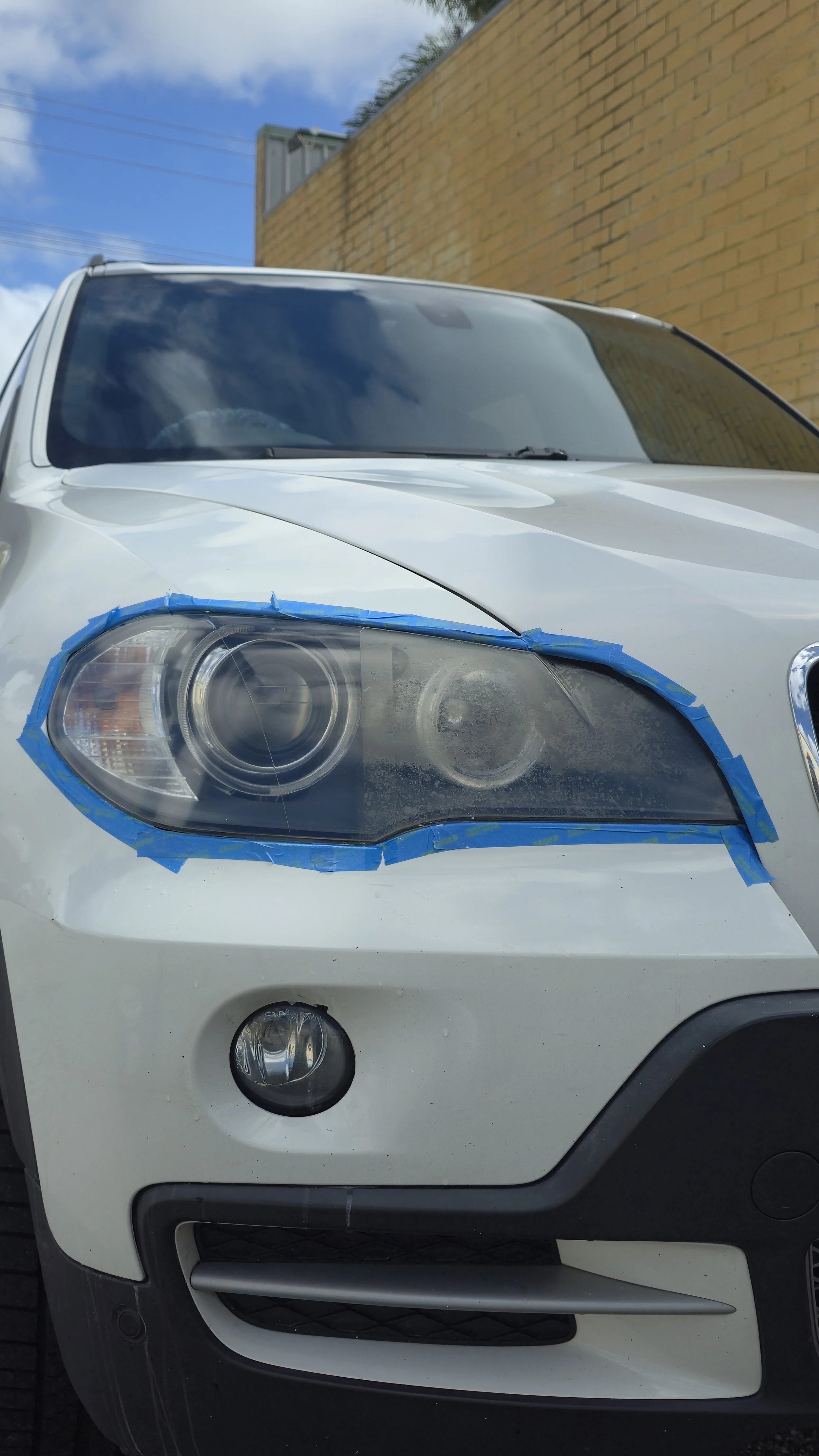 Close-up of a white car with the front left headlight taped with blue painter's tape, parked outdoors near a brick wall.