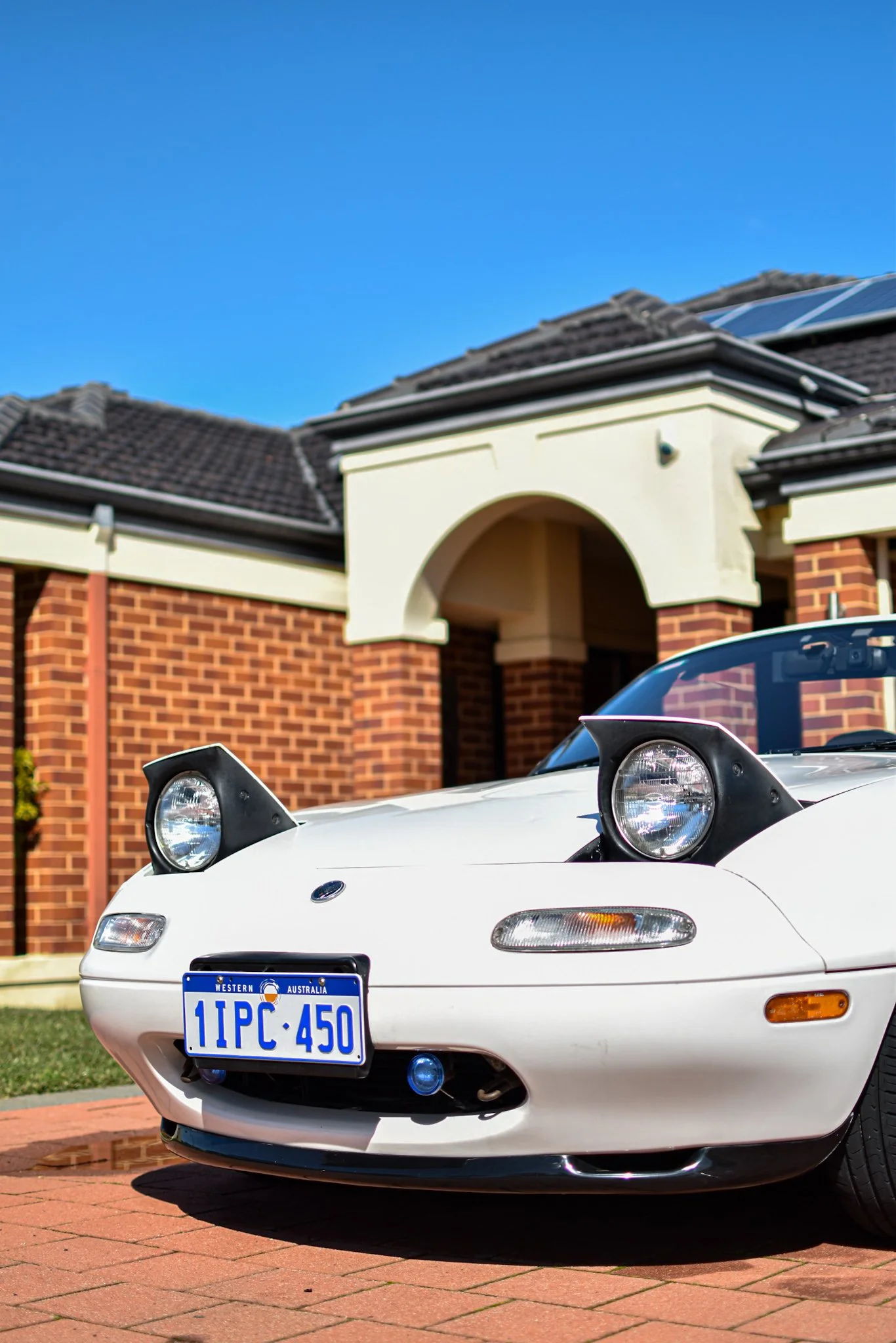 A white sports car parked on a brick driveway in front of a modern brick house with solar panels on the roof and a bright blue sky above.