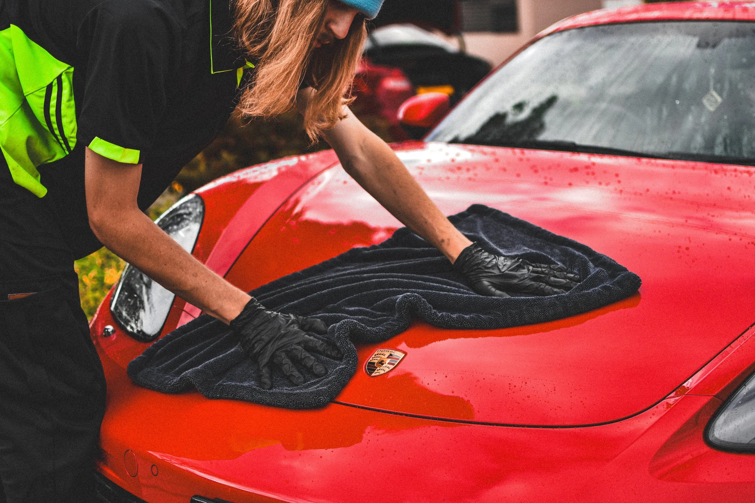 A person in a high-visibility jacket and gloves is using a towel to clean a red Porsche sports car.