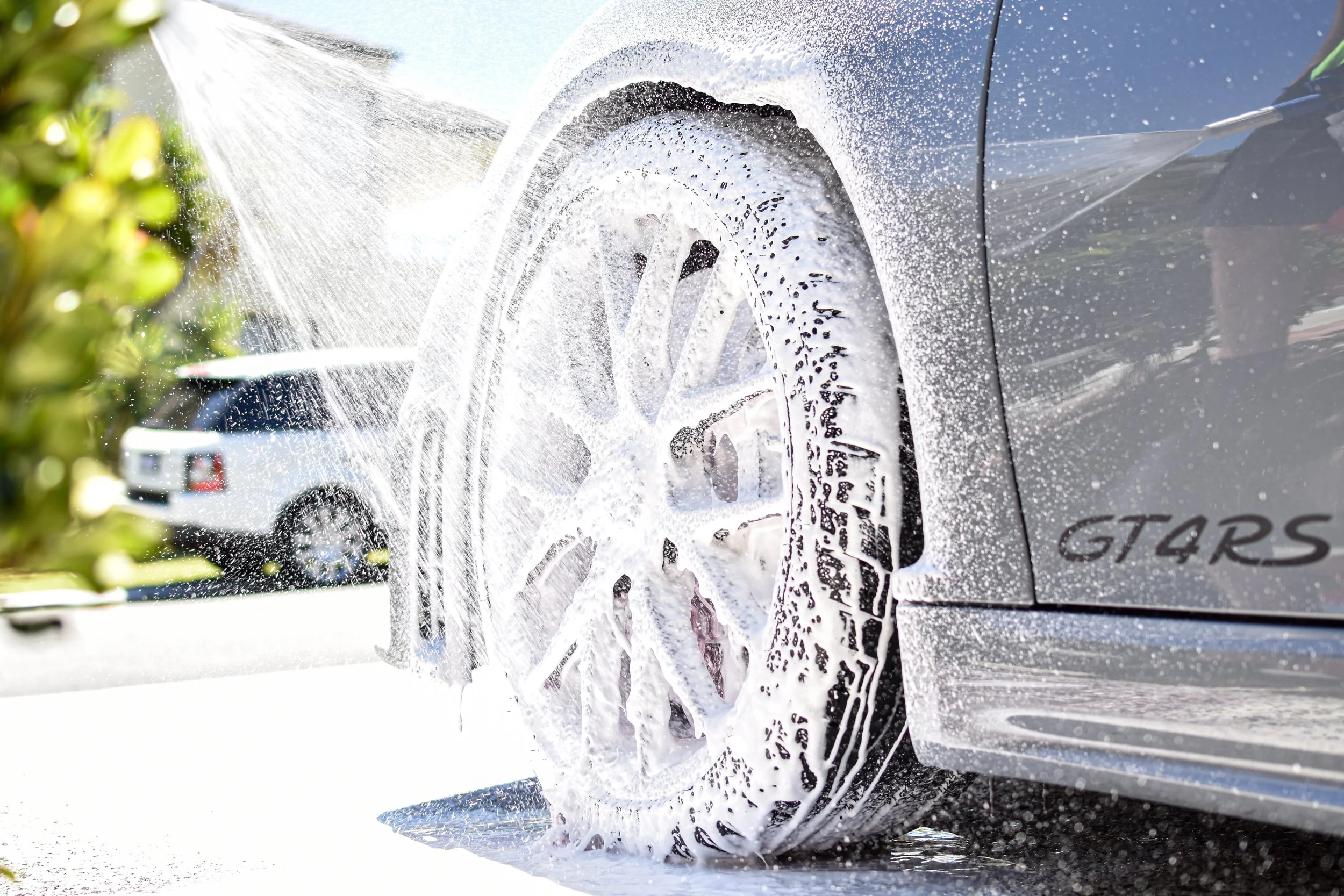 Close-up of a car's wheel being washed with soap and water under pressure, with soap suds covering the tire and rim, and a white vehicle reflected in the car's body.