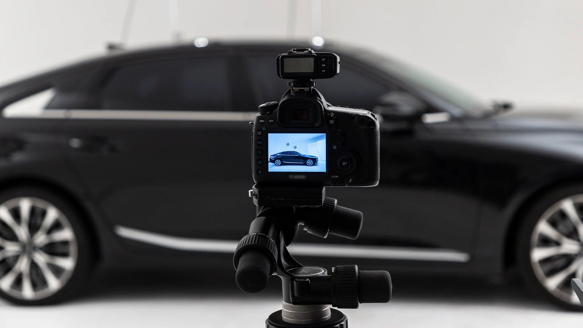 A professional camera mounted on a tripod, capturing a sleek black luxury sedan in a studio setting.