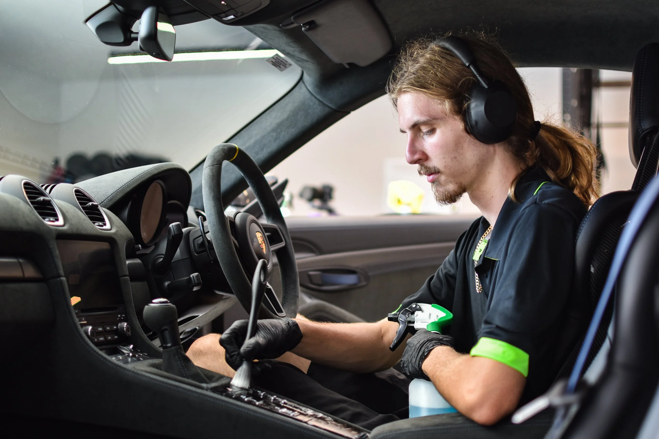 A man in a black shirt and black gloves cleaning the interior of a car, sitting in the driver's seat, with headphones on, using a spray bottle and cloth in a garage or workshop.