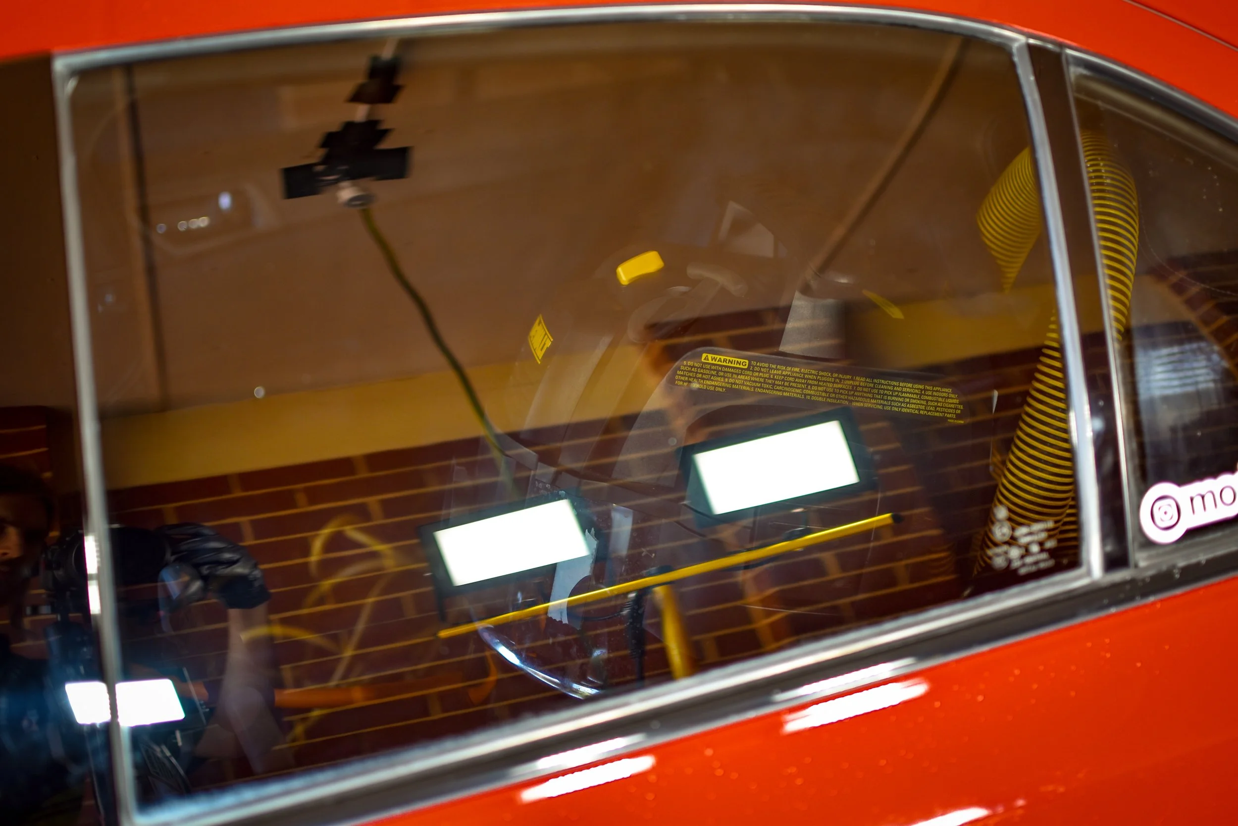 A red vintage car seen through its windshield, with interior yellow-striped seats, yellow tubing on the right side, and two illuminated screens reflected on the glass. A person is reflected taking the photo.