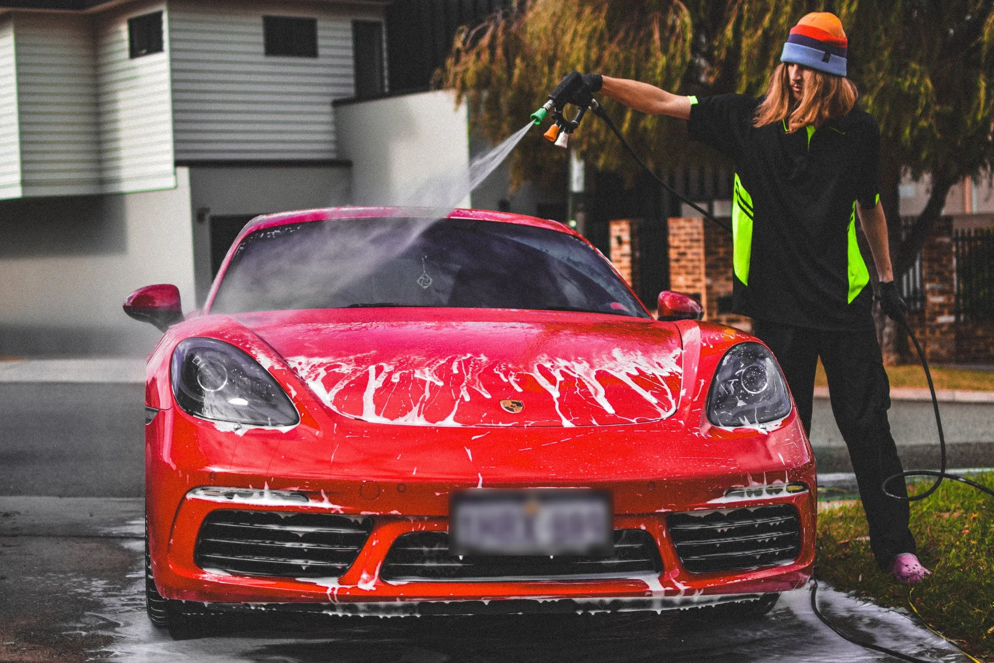 A woman wearing a striped beanie and gloves is using a hose to wash a red Porsche sports car during daytime.