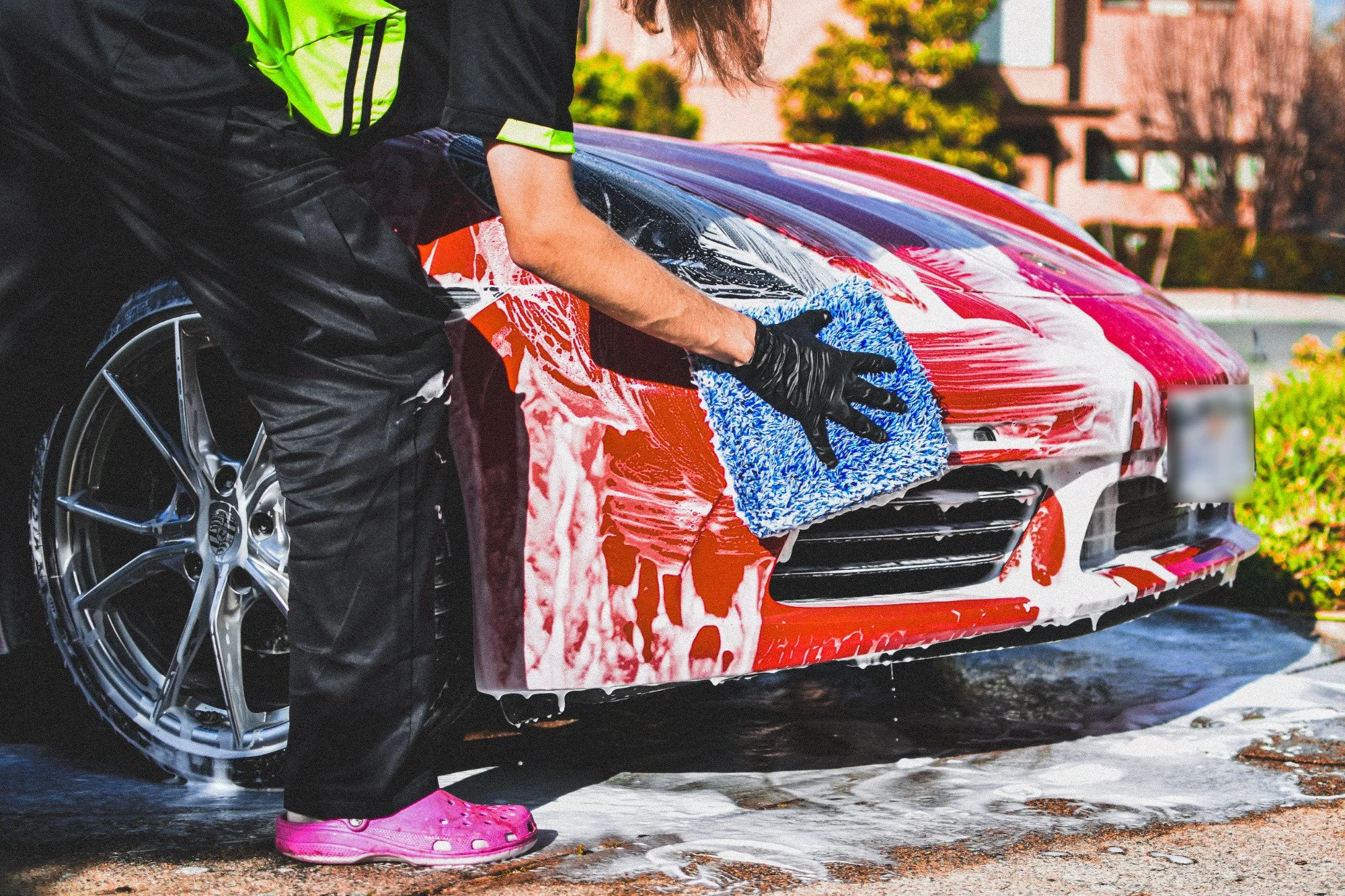 Person washing a red sports car with soap and sponge outdoors.