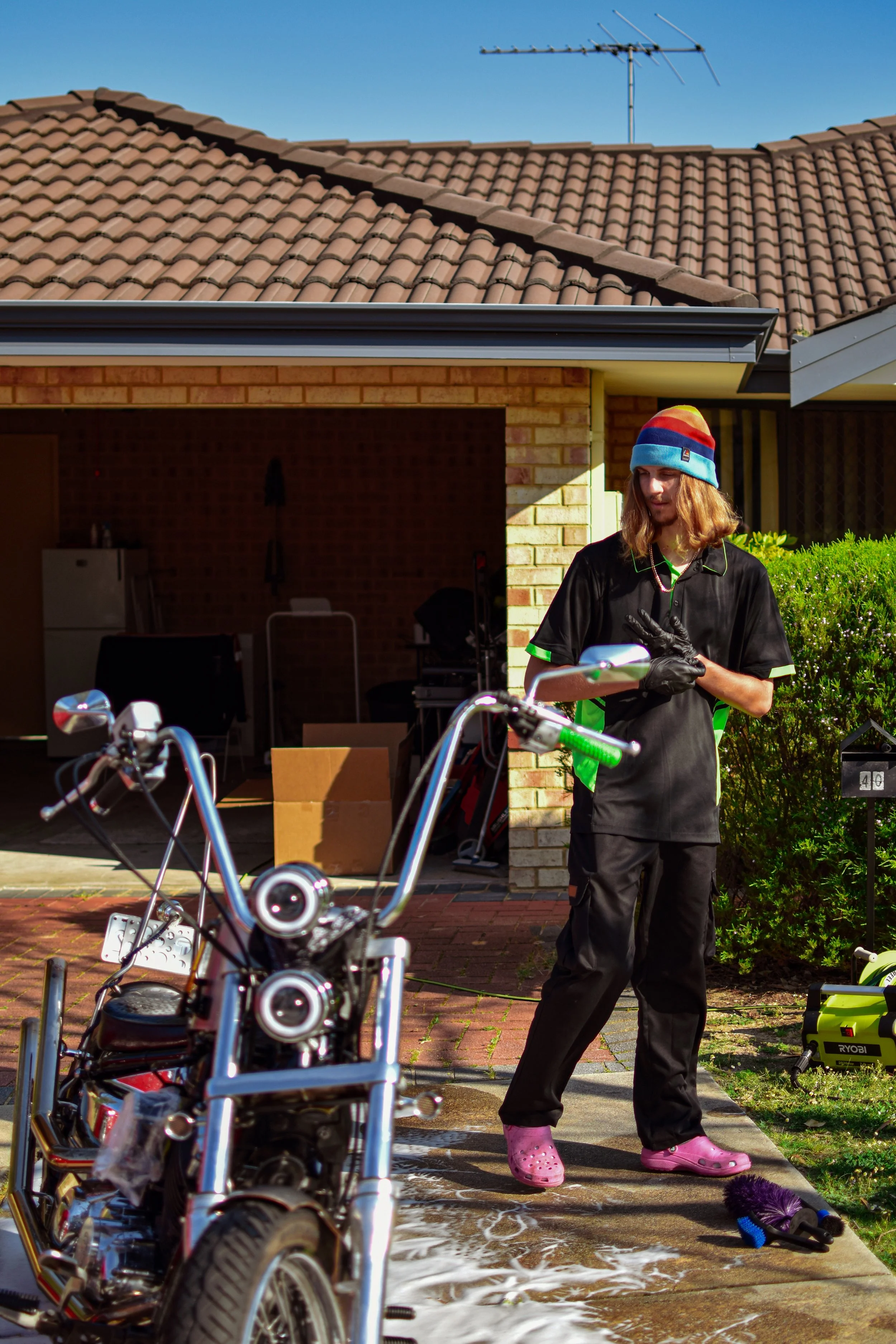 A person in black clothes, pink Crocs, and a colorful beanie is standing outside a house with a motorcycle and soap suds on the ground, possibly washing the motorcycle.
