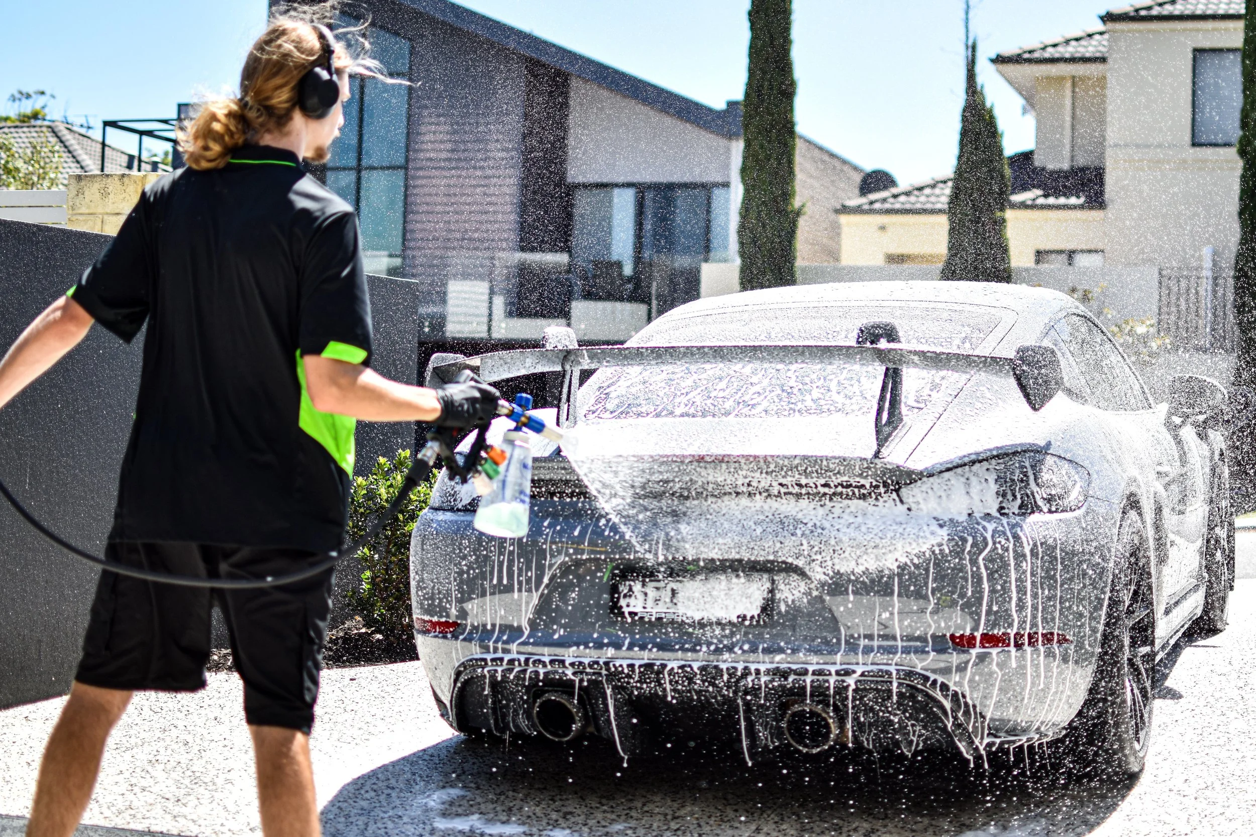 Person wearing sunglasses and black shorts with green accents, using a pressure washer to rinse a silver sports car covered in soap suds outside on a sunny day.