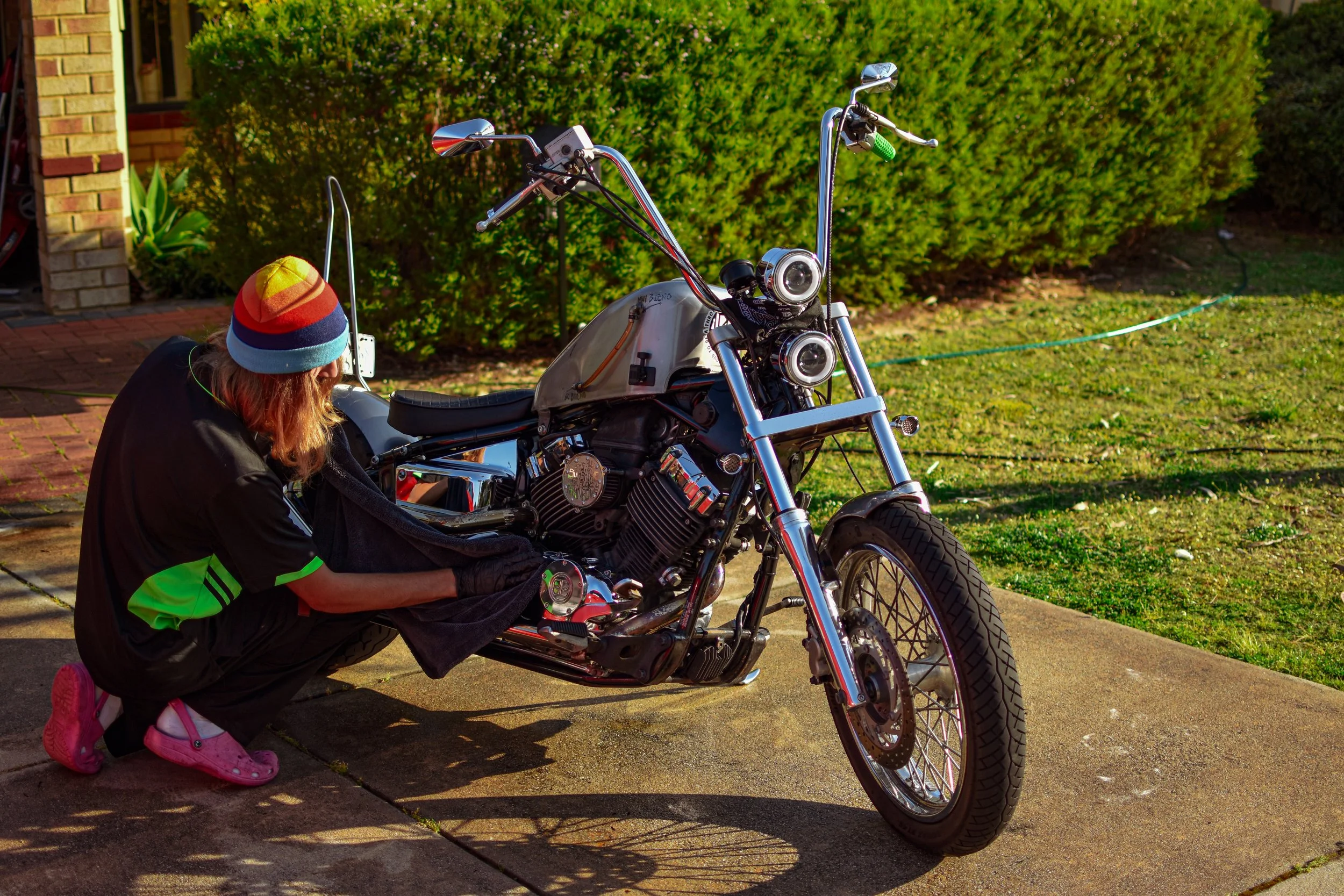 Person crouching next to a motorcycle, cleaning or inspecting it in a driveway with green bushes in the background.