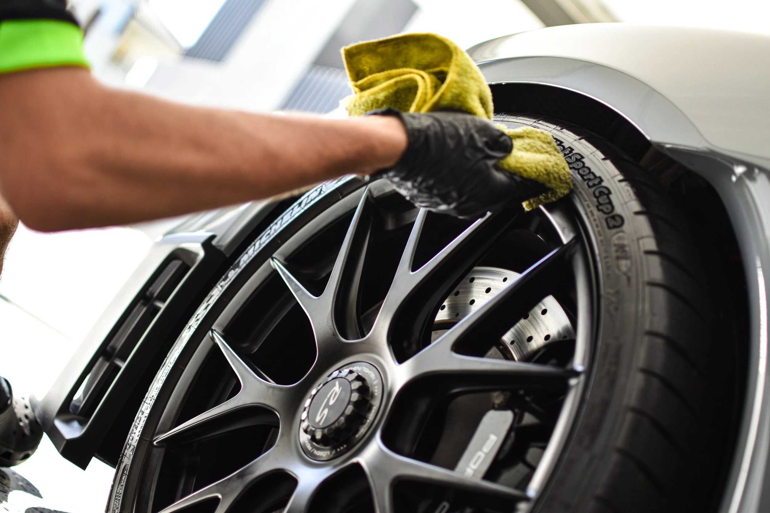 Person cleaning a car wheel with a yellow cloth, wearing a black glove.