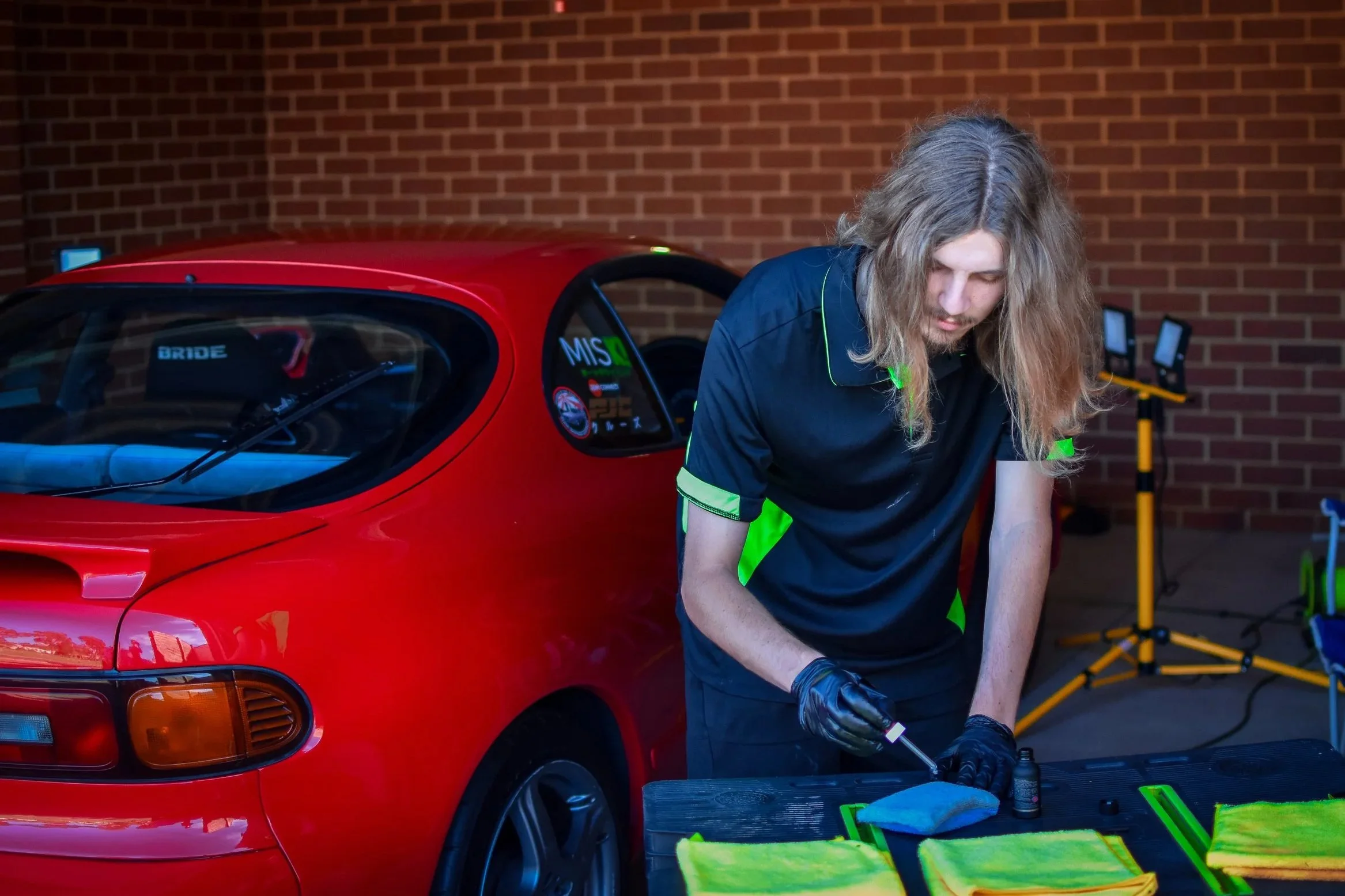 A man with long blond hair wearing a black and neon green polo shirt cleaning a small red race car in a garage or workshop.
