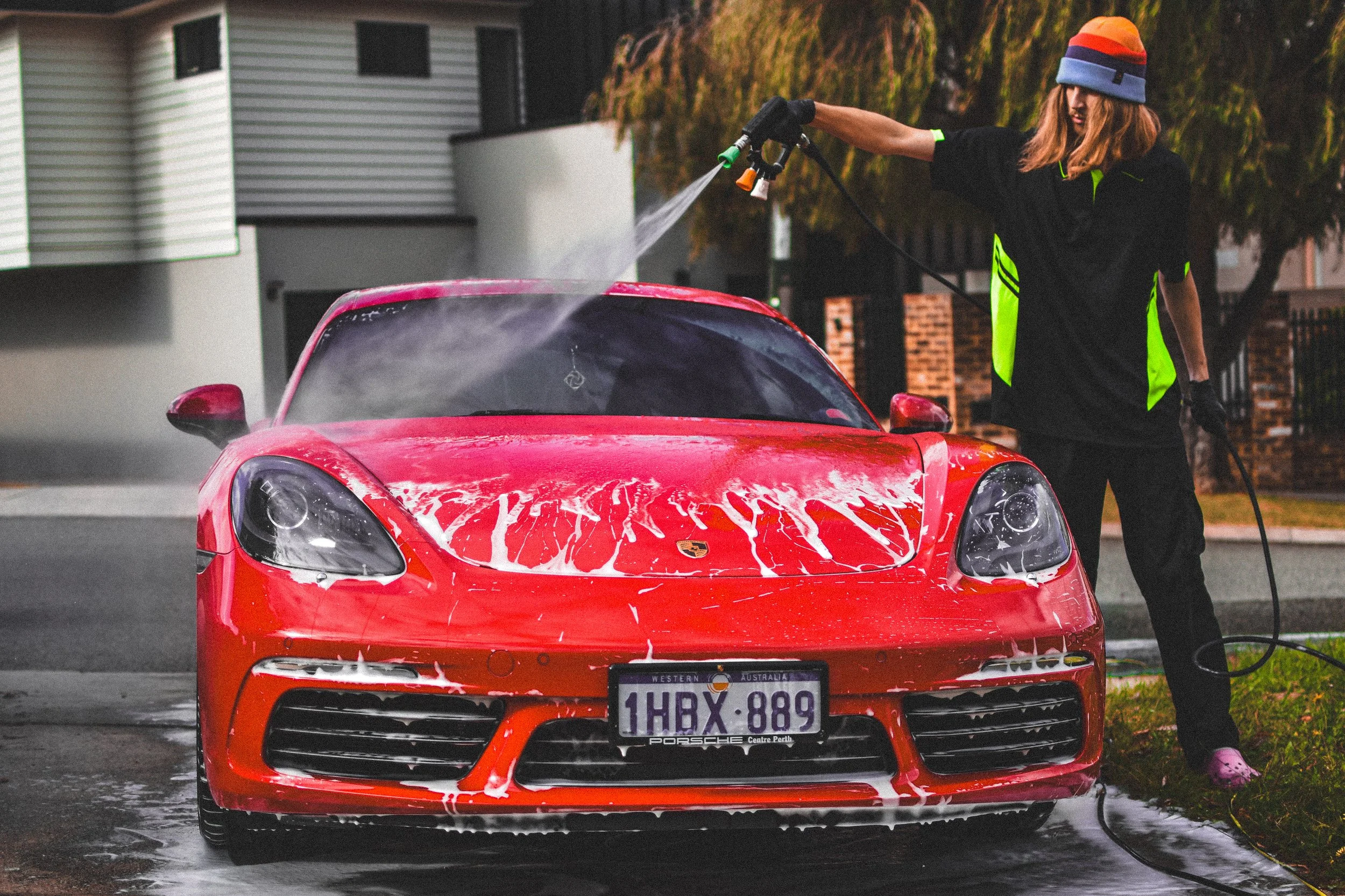 A woman washing a red Porsche sports car with soap and a hose on a driveway of a residential neighborhood.