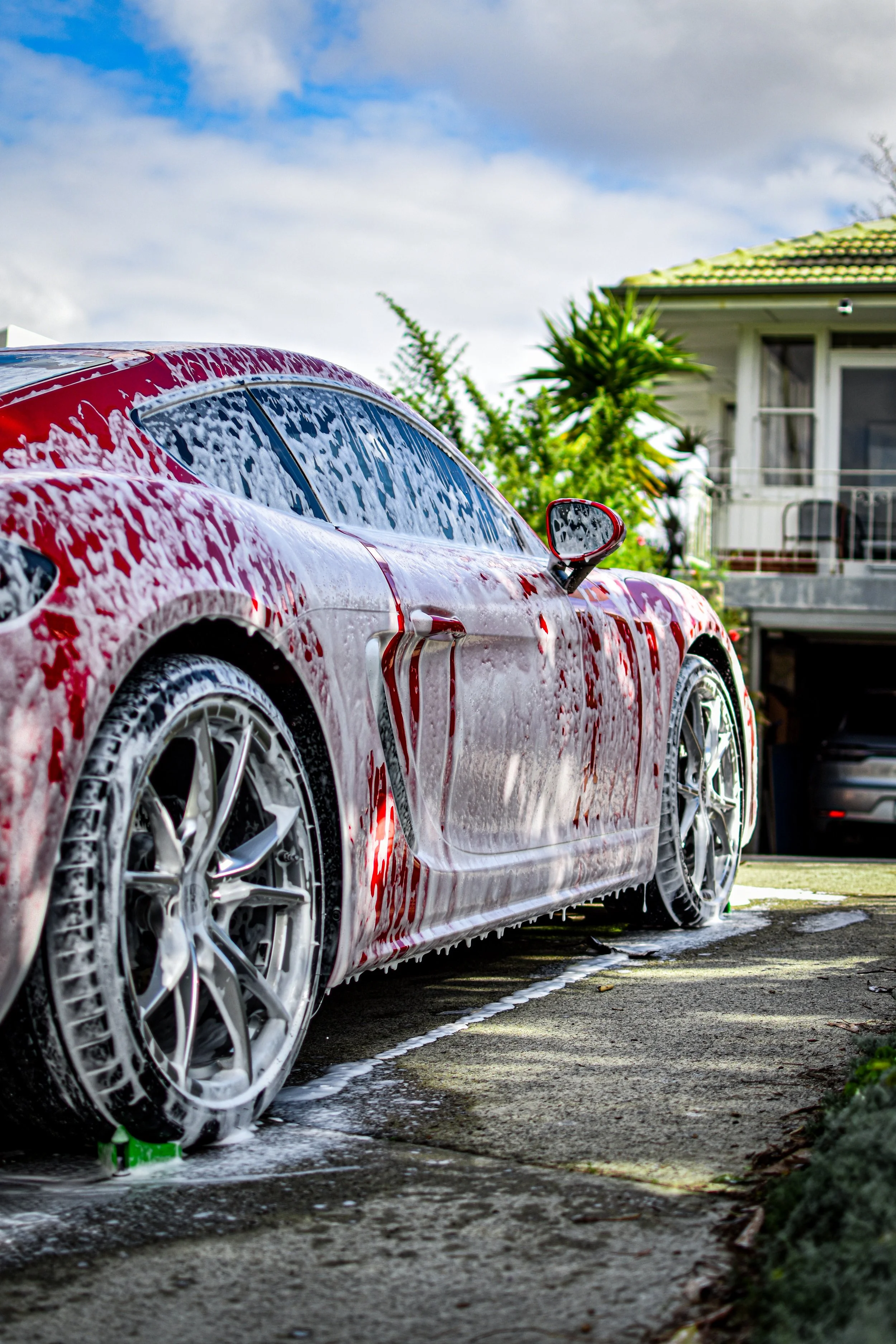 A red sports car covered in soap suds during a wash, with foam dripping down its sides, parked on a driveway with a house and trees in the background.