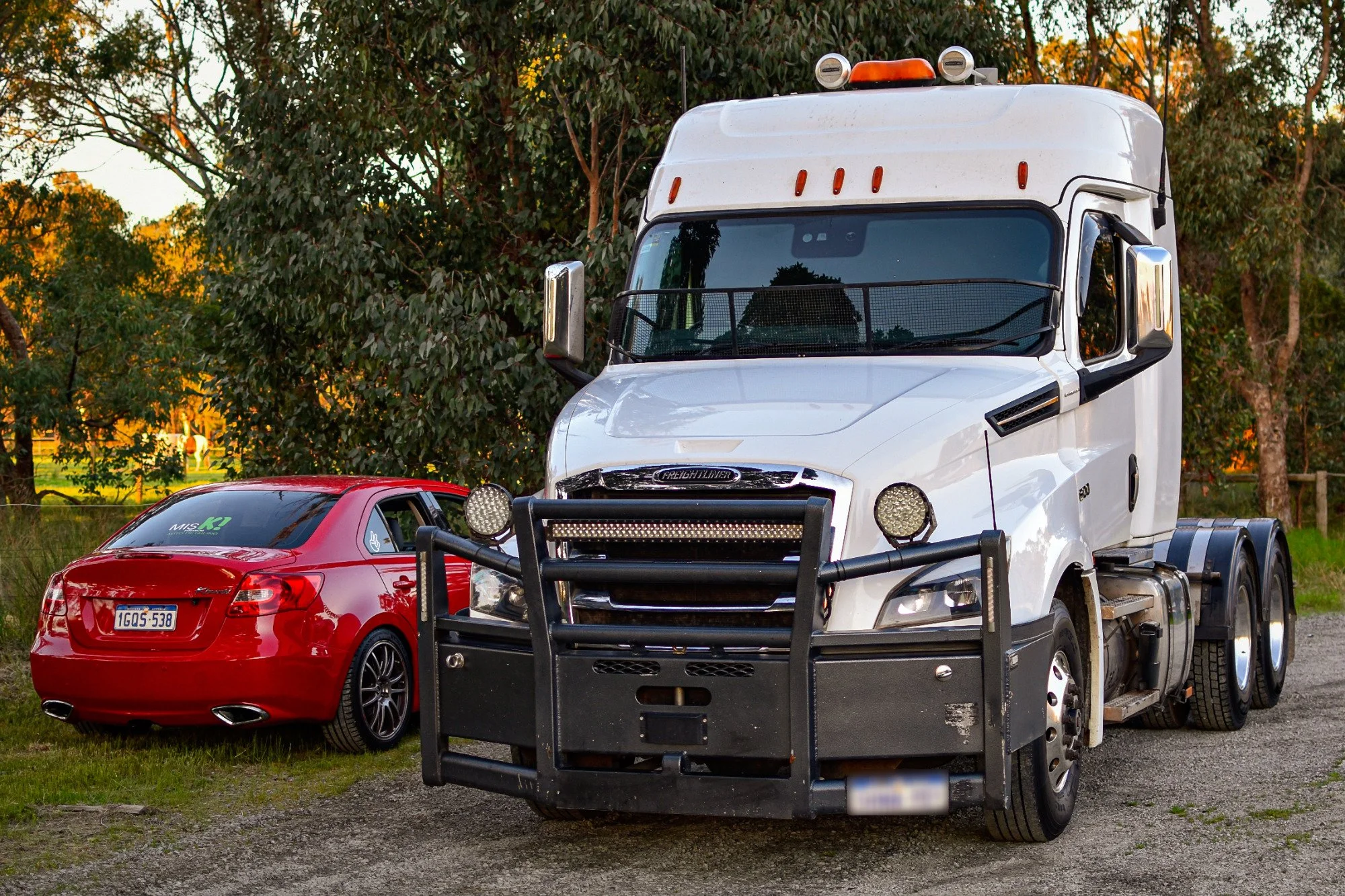 A white semi-truck with a police-style grille guard parked on a gravel lot next to a red sedan, with trees and a sunset in the background.
