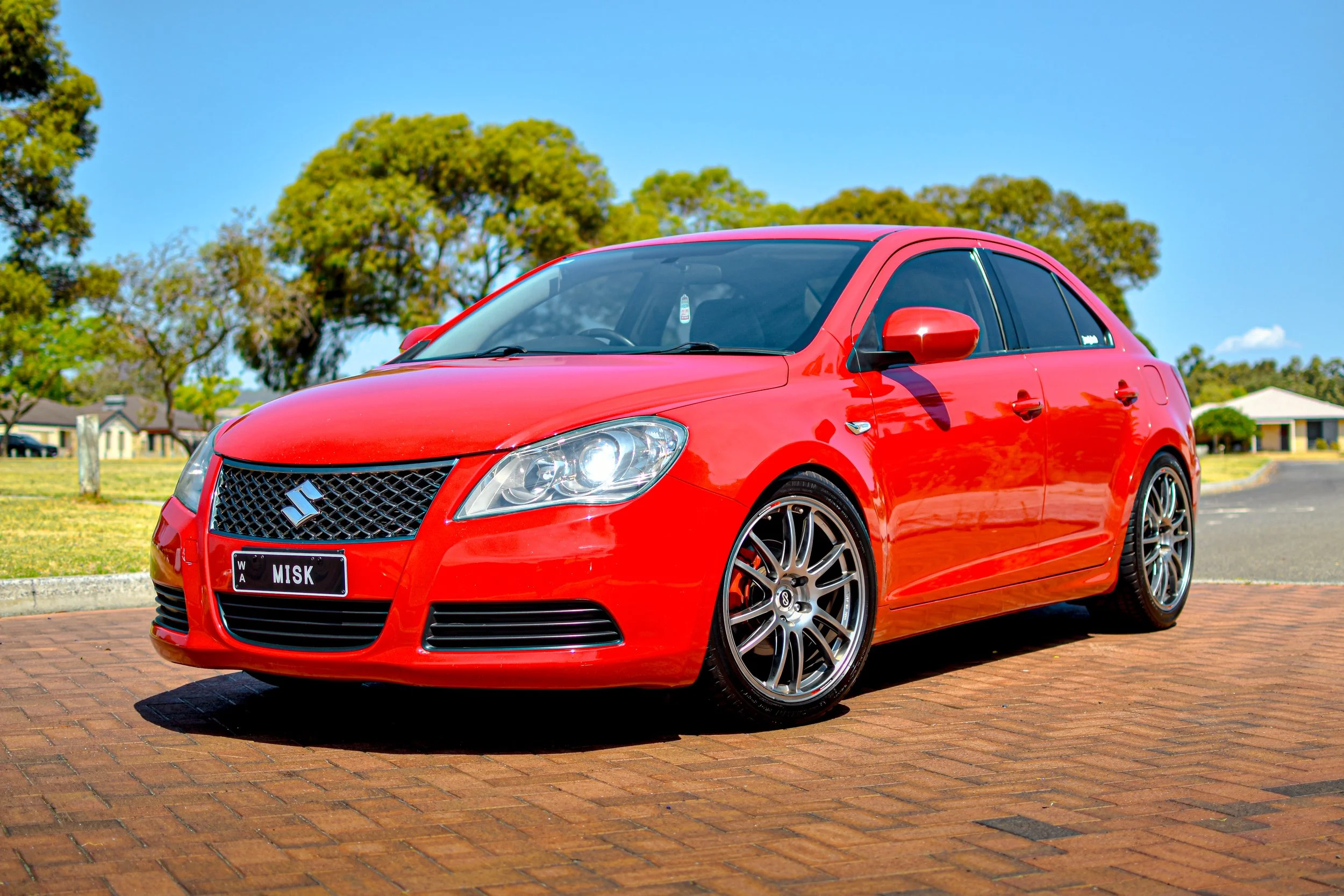 Red sedan parked on a brick driveway with trees and houses in the background under a clear blue sky.