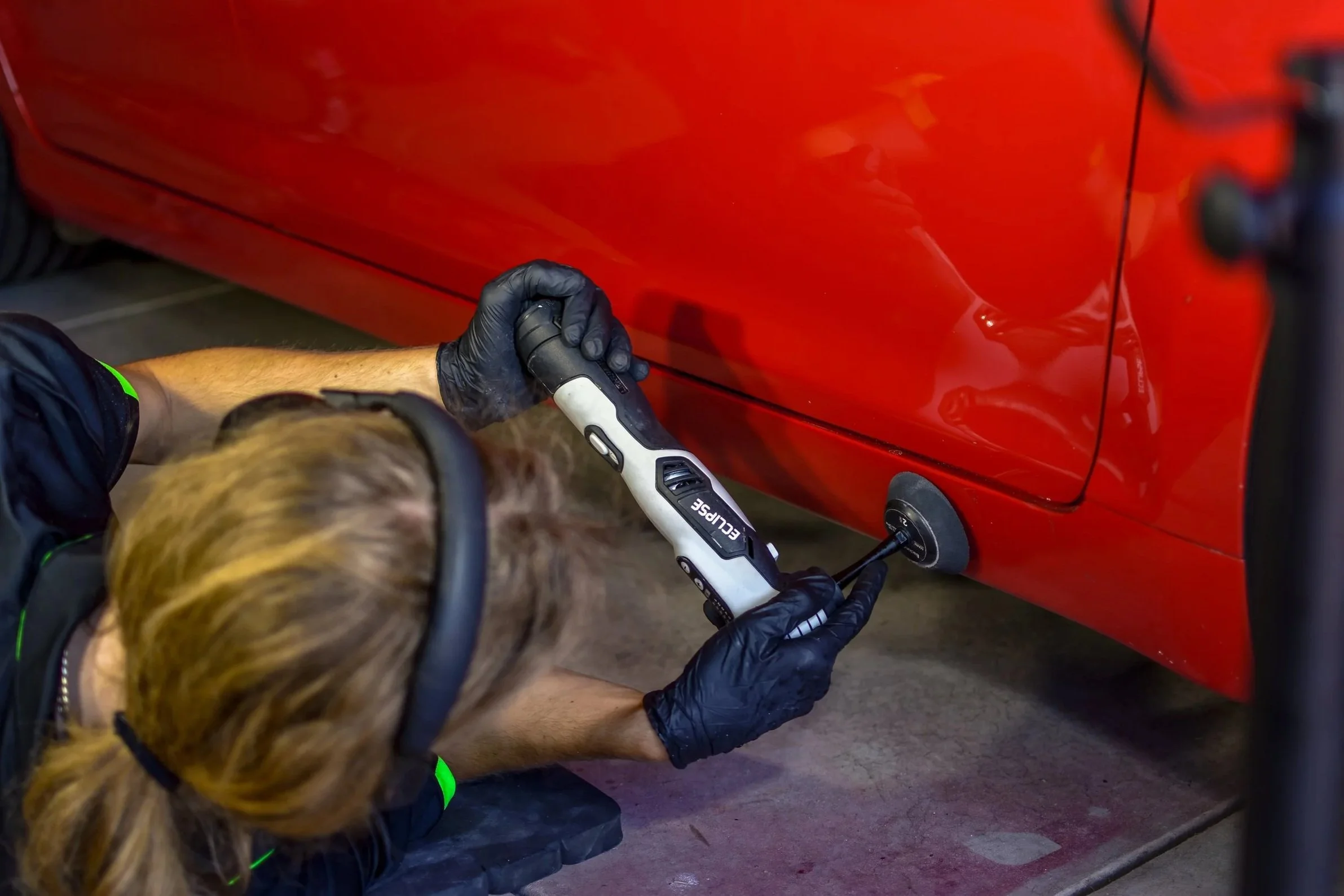 A person wearing black gloves and headphones uses a cordless electric tool to work on the underside of a red car.