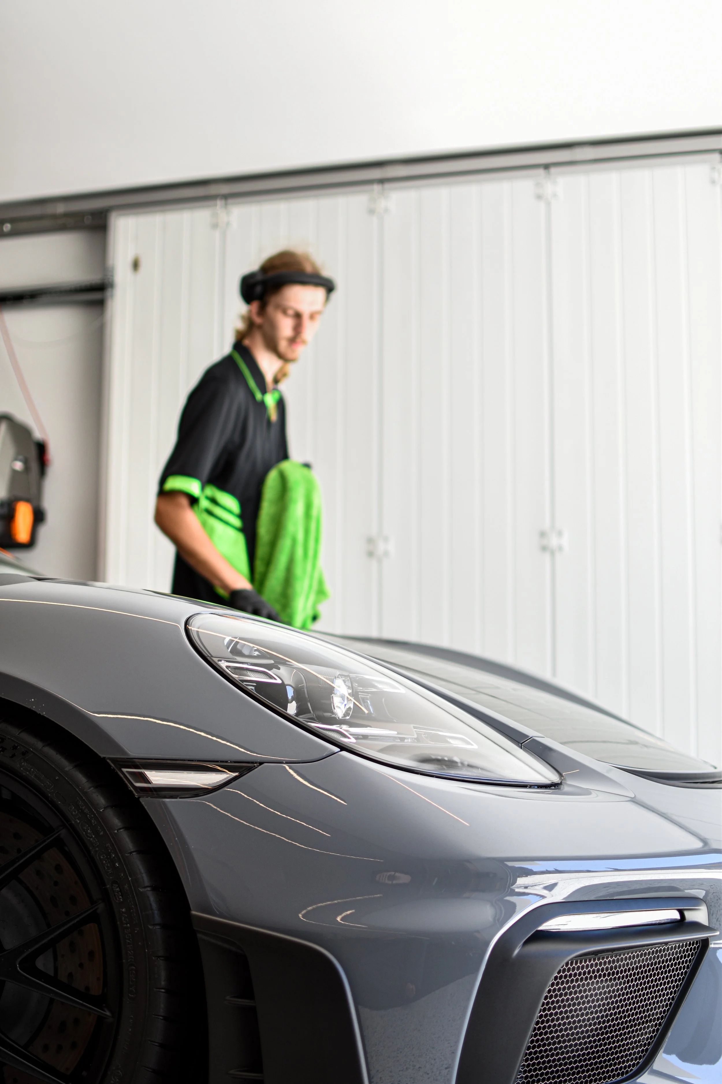 A person in a garage with a large gray sports car, wearing headphones, black and green clothing, and a green towel over their shoulder, with white garage doors in the background.
