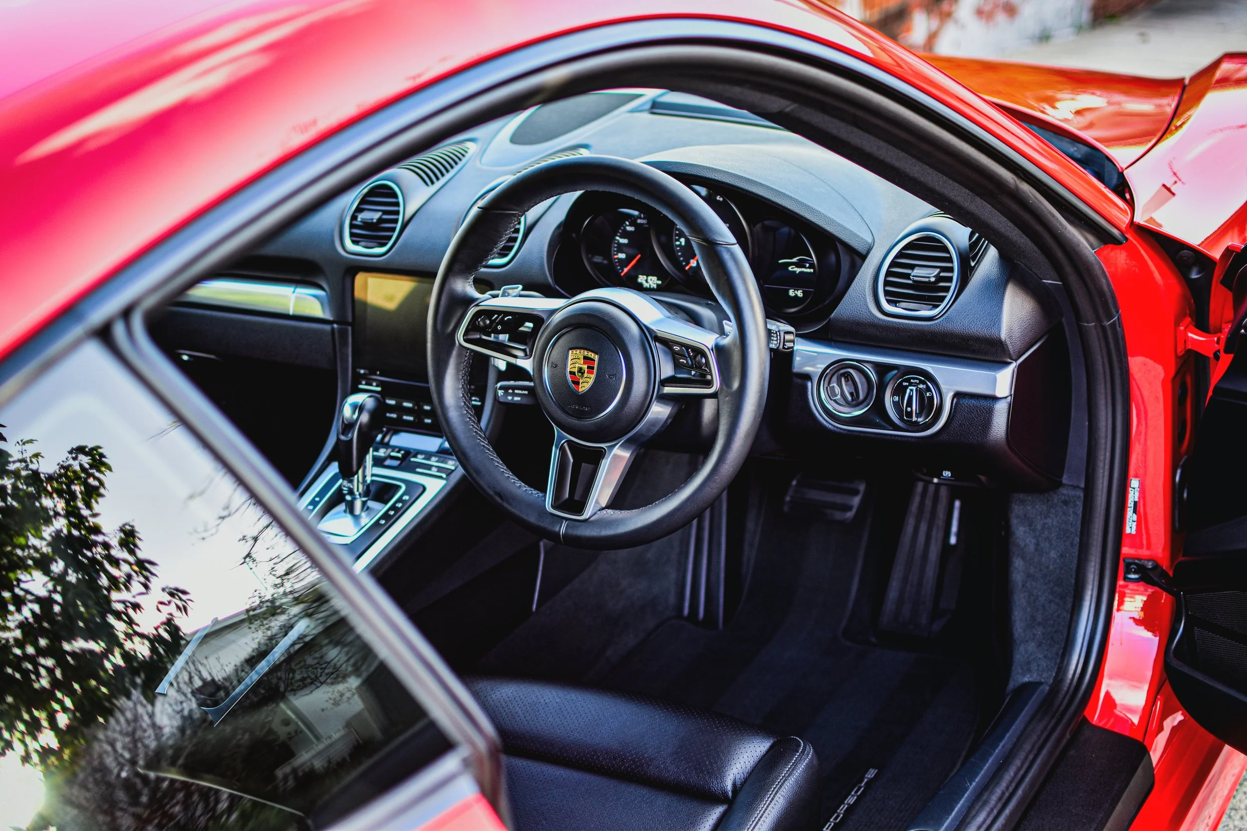 Interior of a red Porsche sports car, showing the steering wheel with the Porsche emblem, dashboard, gear shift, and black leather seat.