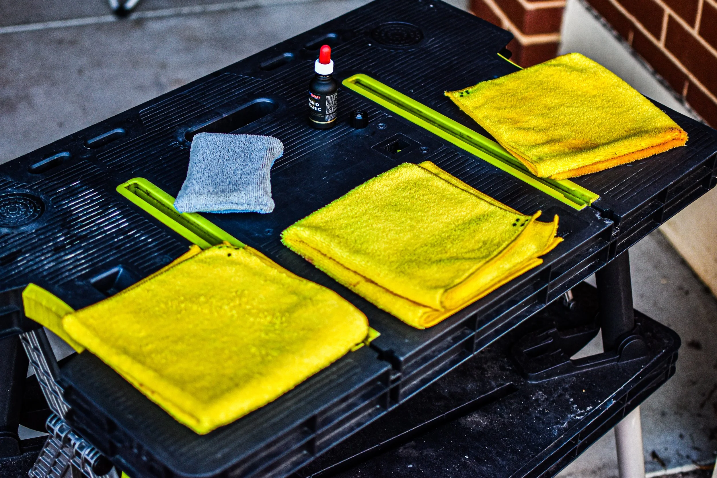 A black workbench with three folded yellow cleaning towels, a bottle of liquid glue, a gray cleaning cloth, and two yellow-handled tools, likely for working with vinyl or similar material.