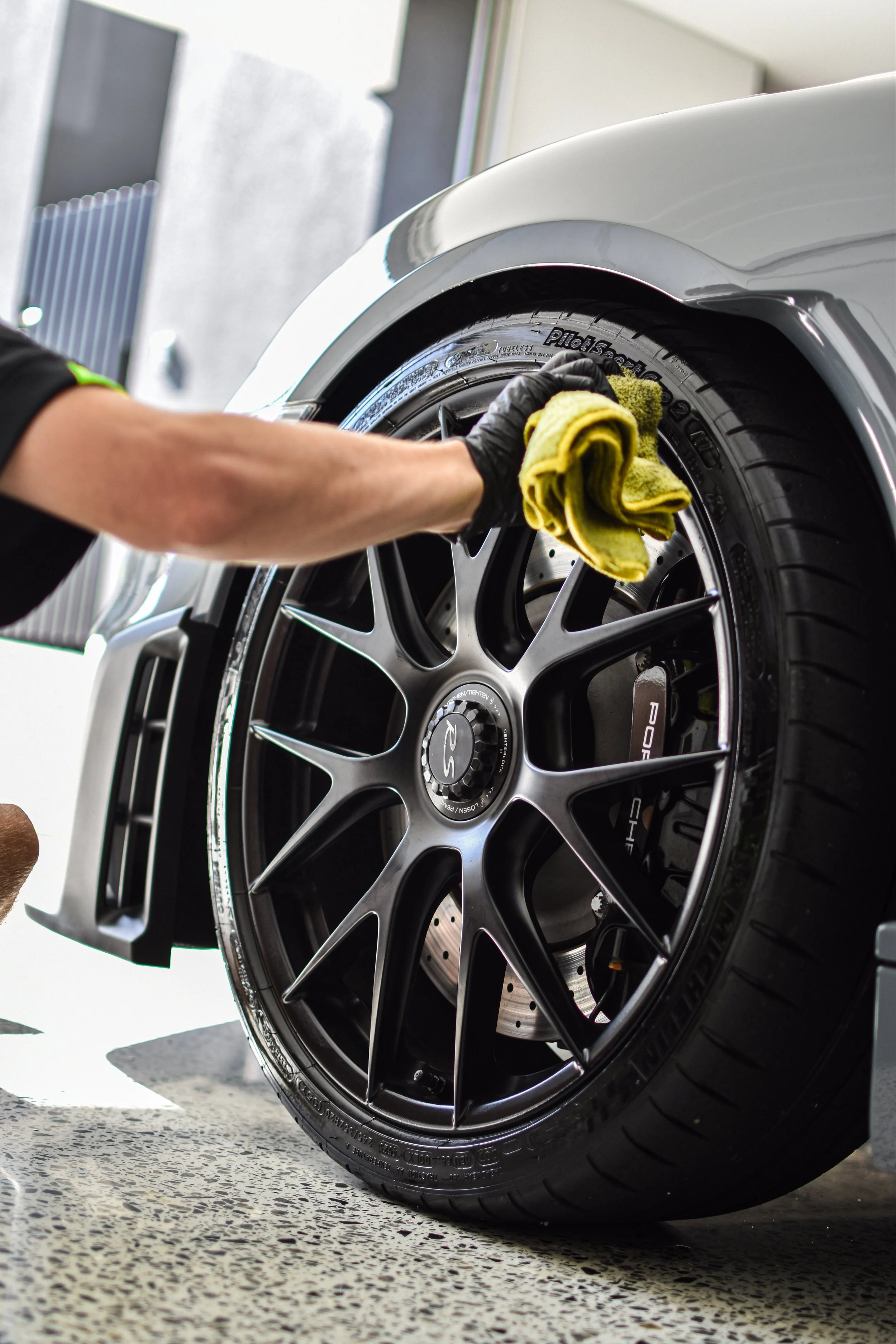 A person wearing a black glove and a black shirt wipes the rim of a black alloy wheel with a yellow towel on a silver sports car.