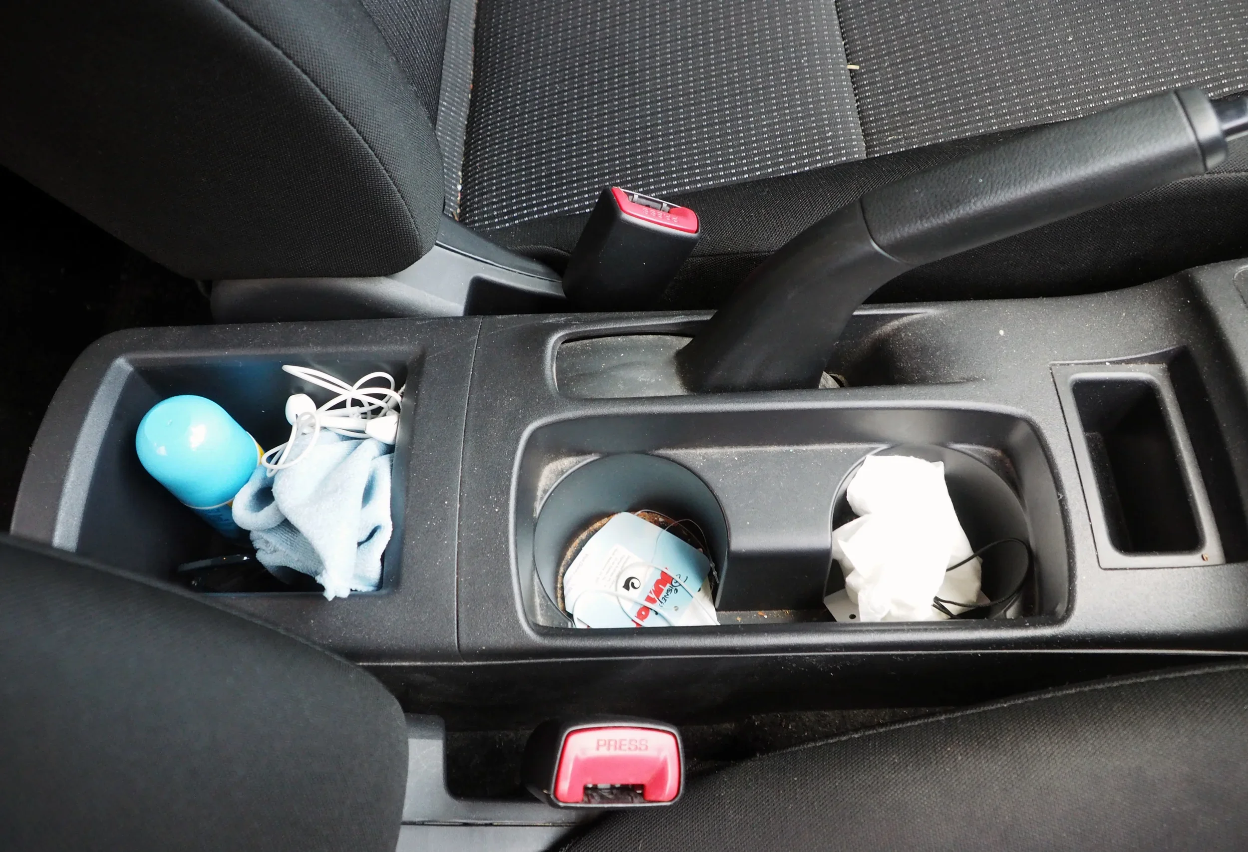 Car interior's center console with clutter, including a blue water bottle, earphones, a box of tissues, and an used face mask.