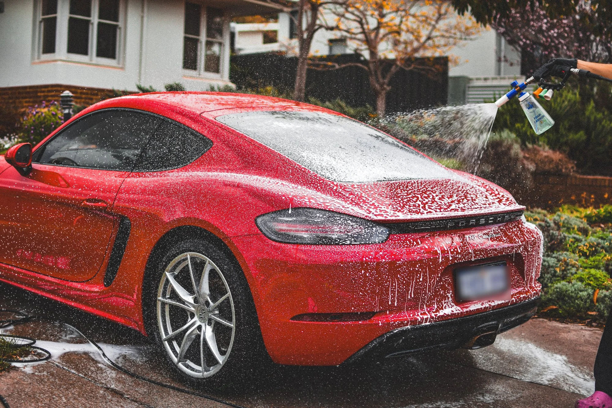 Red sports car being washed with soap and water spray in a residential driveway with a house and trees in the background.