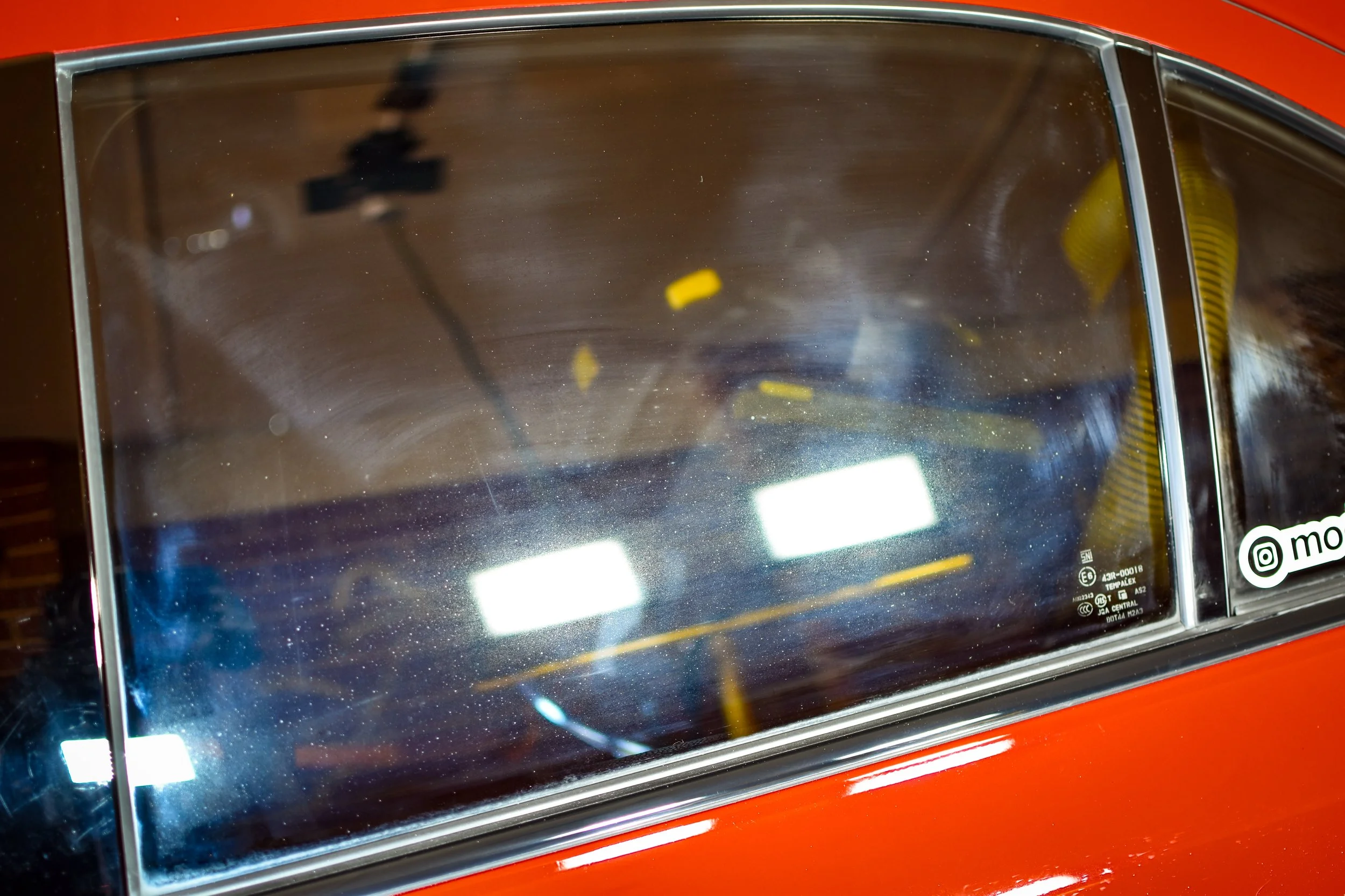 Close-up of the front windshield of a red vintage car, showing smudges, dust, and some yellow leaves inside.