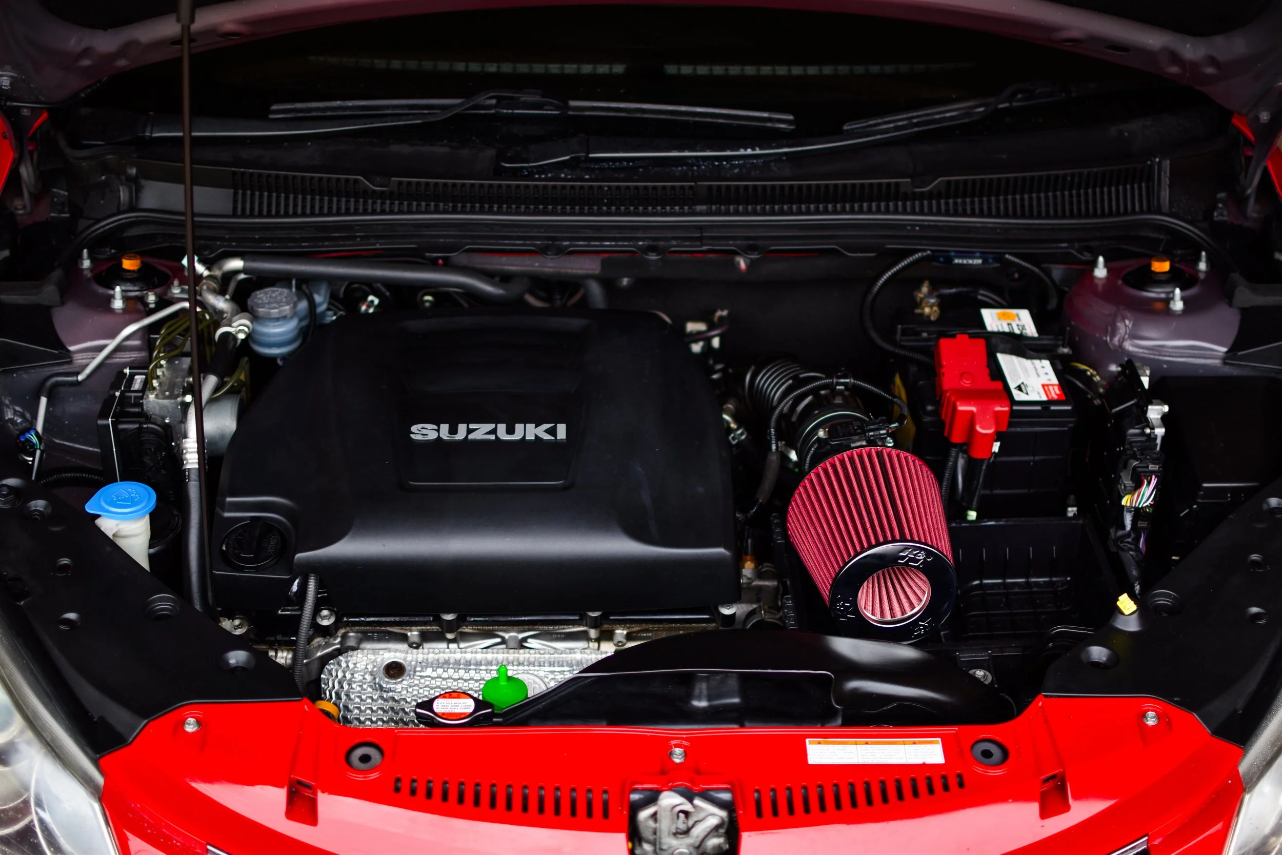 Under the hood of a Suzuki car, showing the engine with a black cover labeled 'Suzuki,' a red air filter, a red battery, and various engine components and hoses.