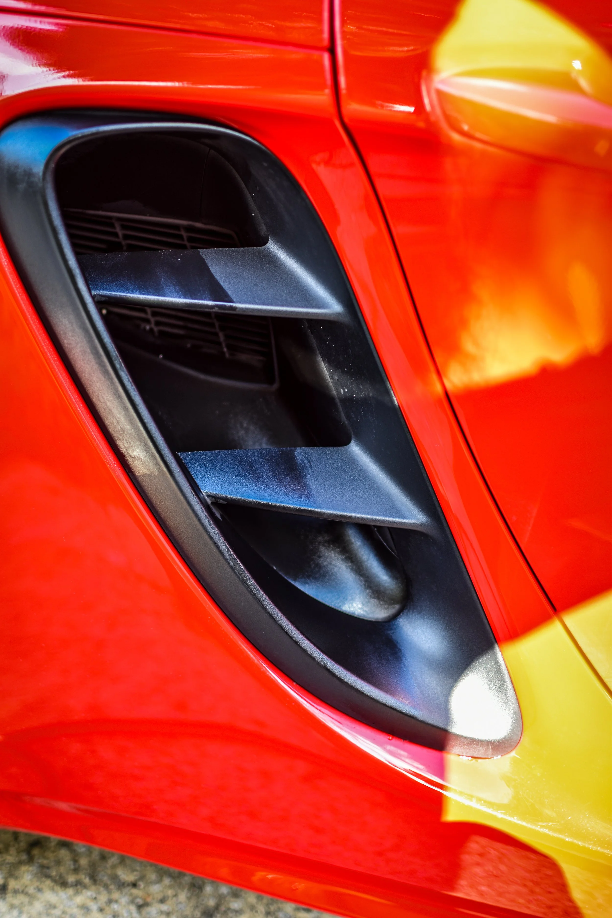 Close-up of a red sports car's front end, featuring a black air vent and part of the yellow headlight.