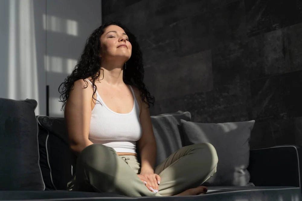 Woman sitting cross-legged on a sofa, relaxing with eyes closed, in a room with natural light and a dark wall behind her.