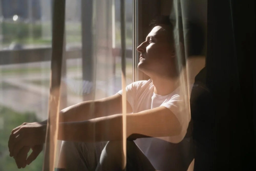 Person sitting by a window, enjoying sunlight with eyes closed.