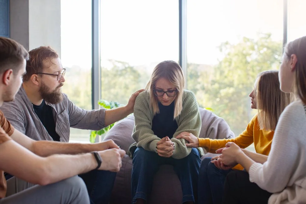 A woman sitting on a couch with her head bowed and eyes closed, surrounded by five people holding her shoulders and hands, in a supportive group therapy or emotional support setting.