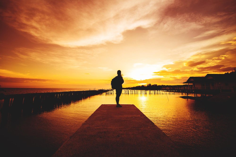 Person standing on a dock by the water during sunset, with colorful sky and calm water reflecting the sunset.