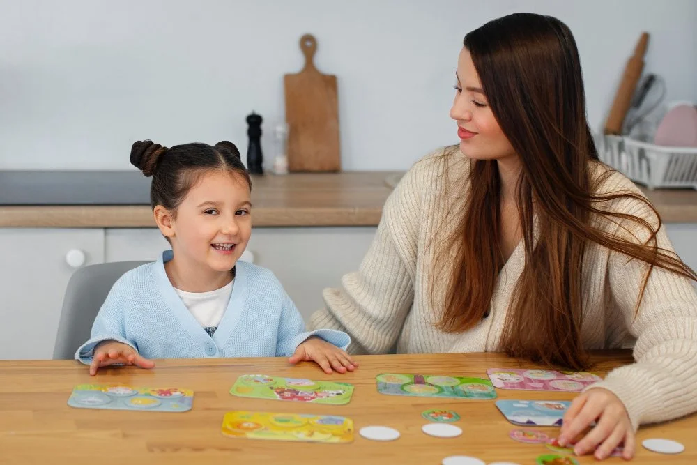 A woman and a young girl playing a card game at a wooden table in a bright kitchen.