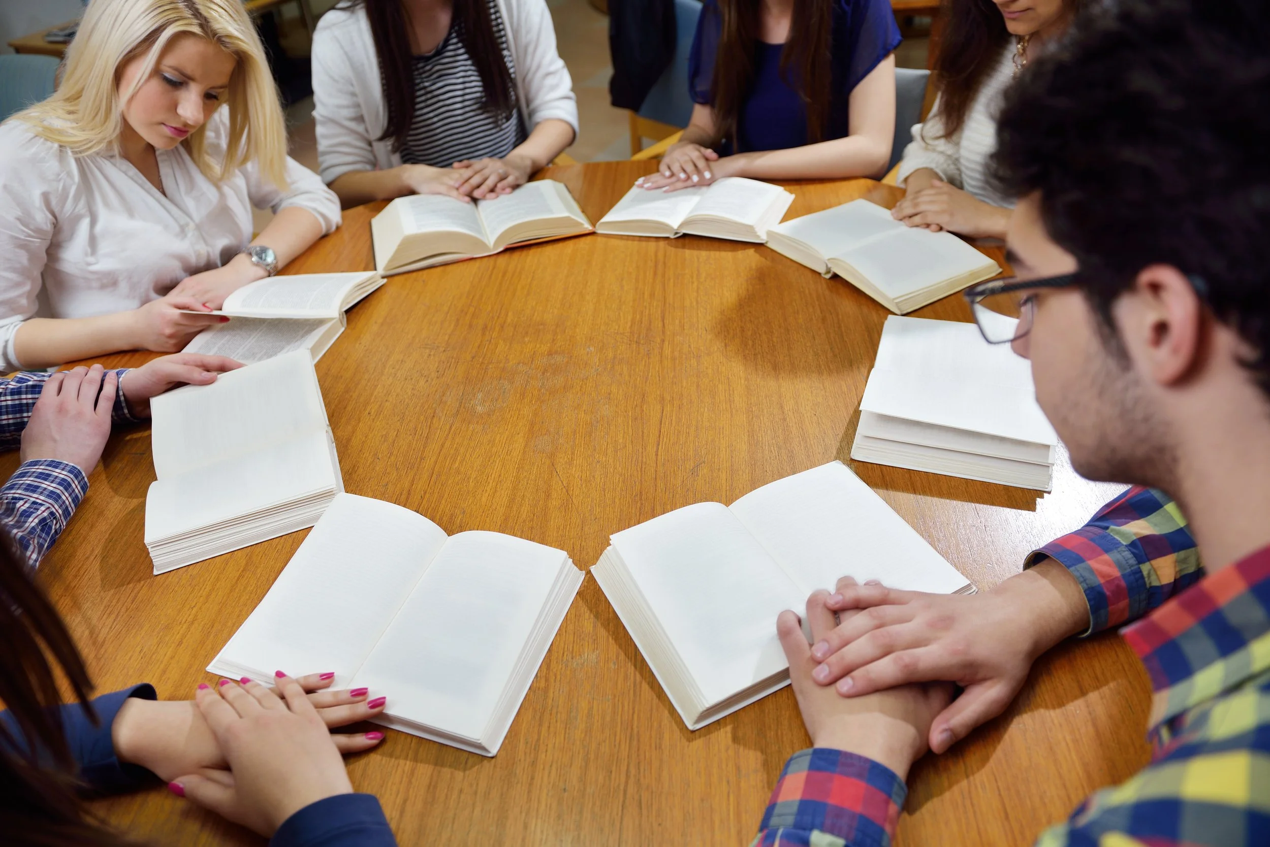 Group of people sitting around a circular wooden table, reading open books, in a study or meeting setting.
