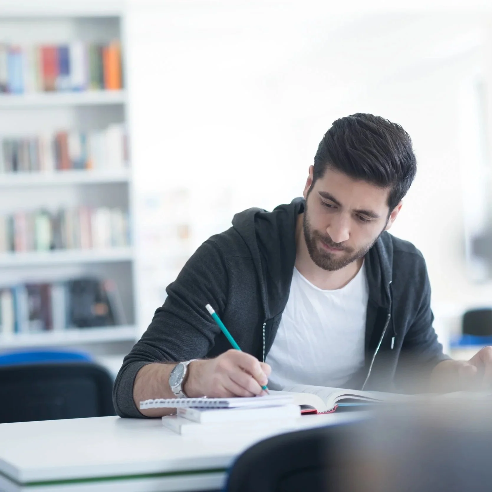 A young man with dark hair and a beard sitting at a desk, writing in a notebook with a blue pen, in a well-lit room with bookshelves in the background.