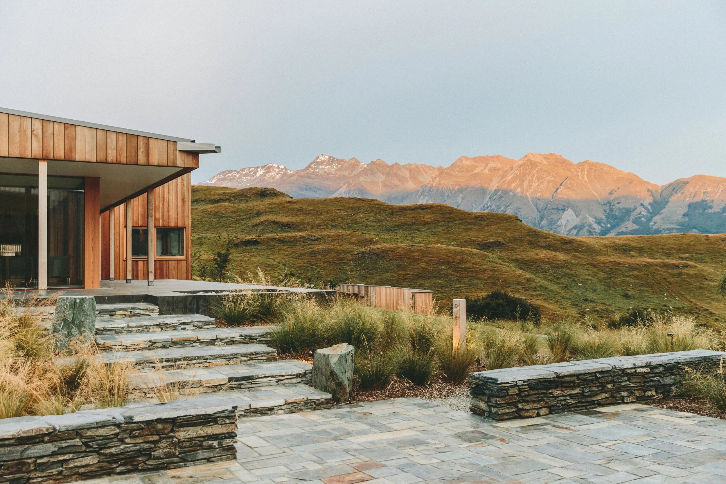 Modern house with wooden exterior, stone steps, and mountain landscape in the background at sunset.
