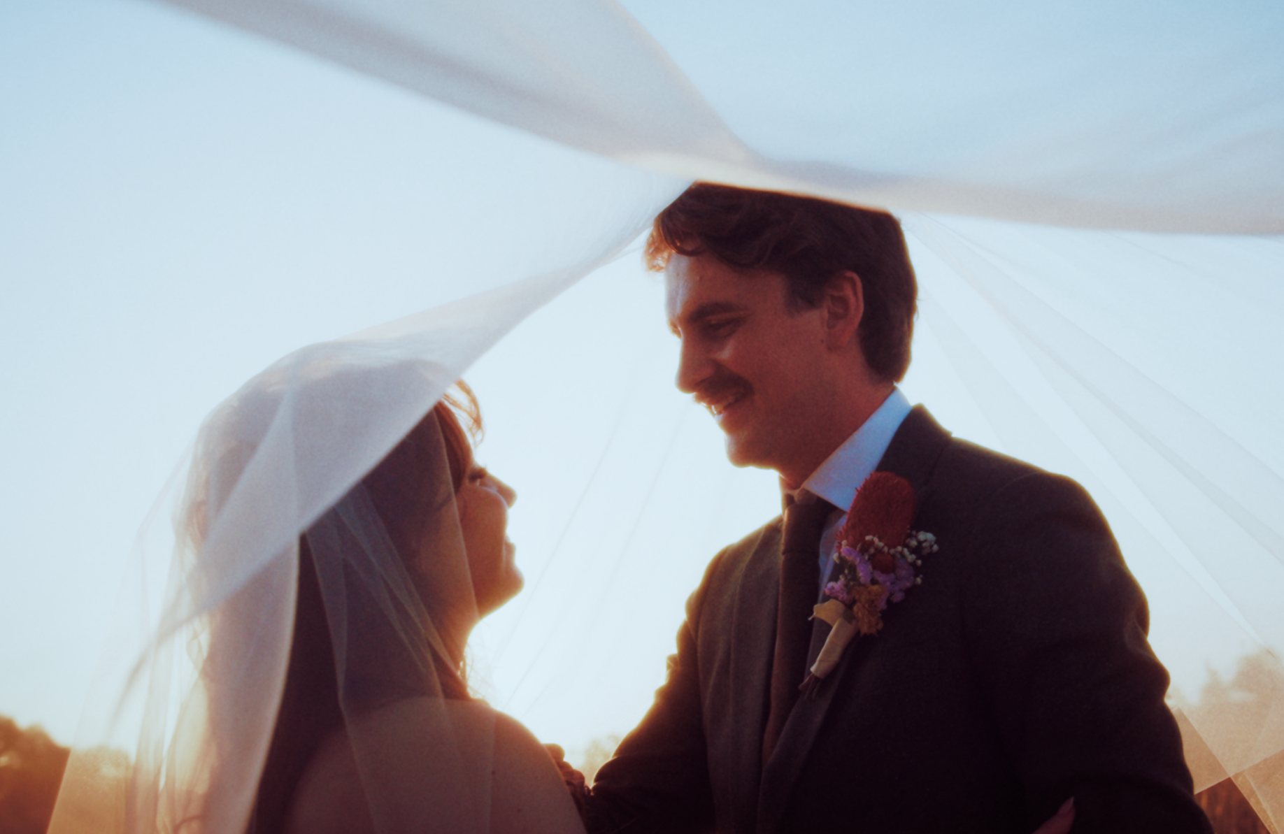 A bride and groom smiling at each other under a flowing wedding veil outdoors during sunset.