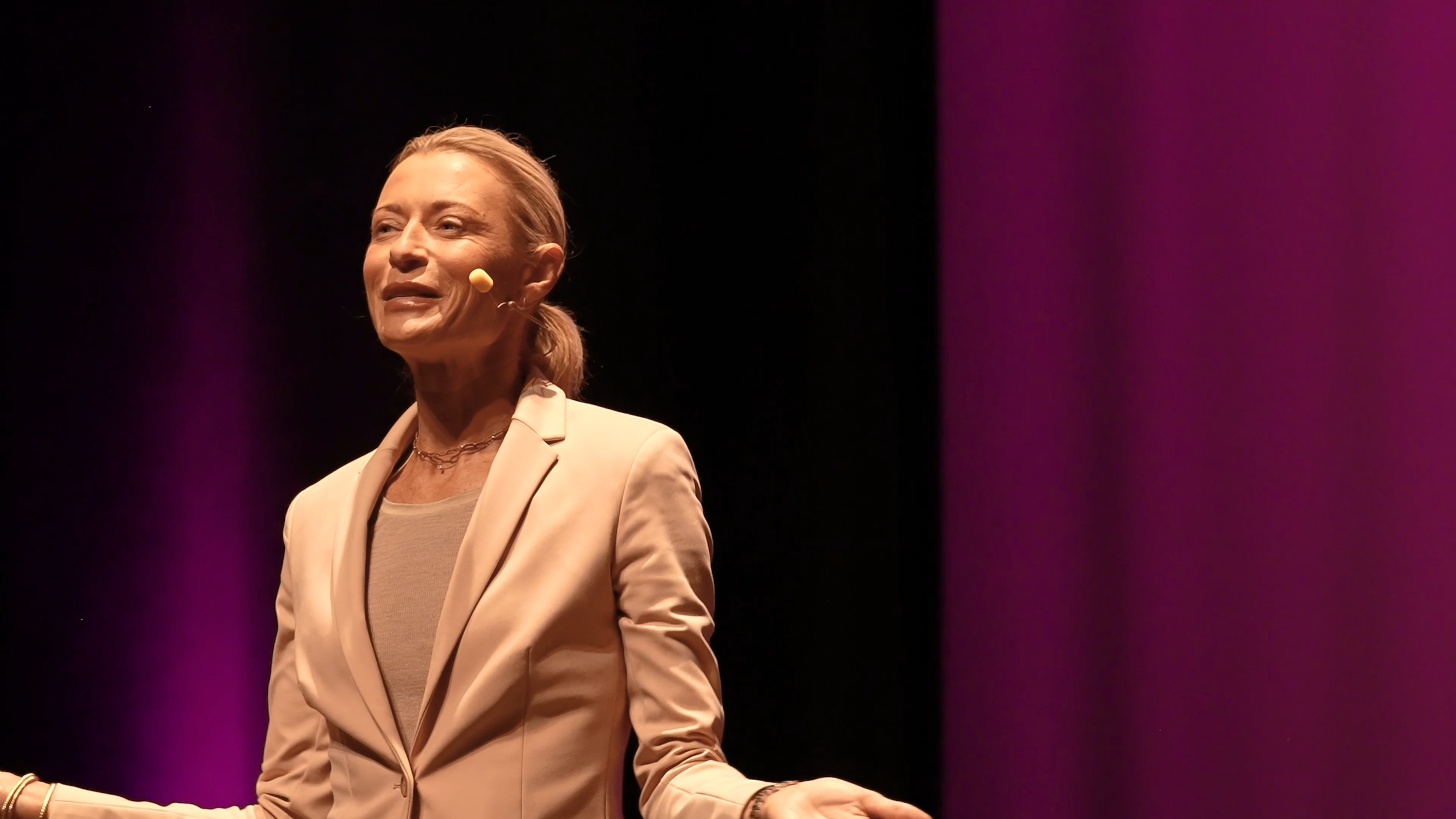 A middle-aged woman with blonde hair tied back, wearing a light beige blazer and a beige top, standing on stage with a black background and purple lighting, speaking into a head-worn microphone.