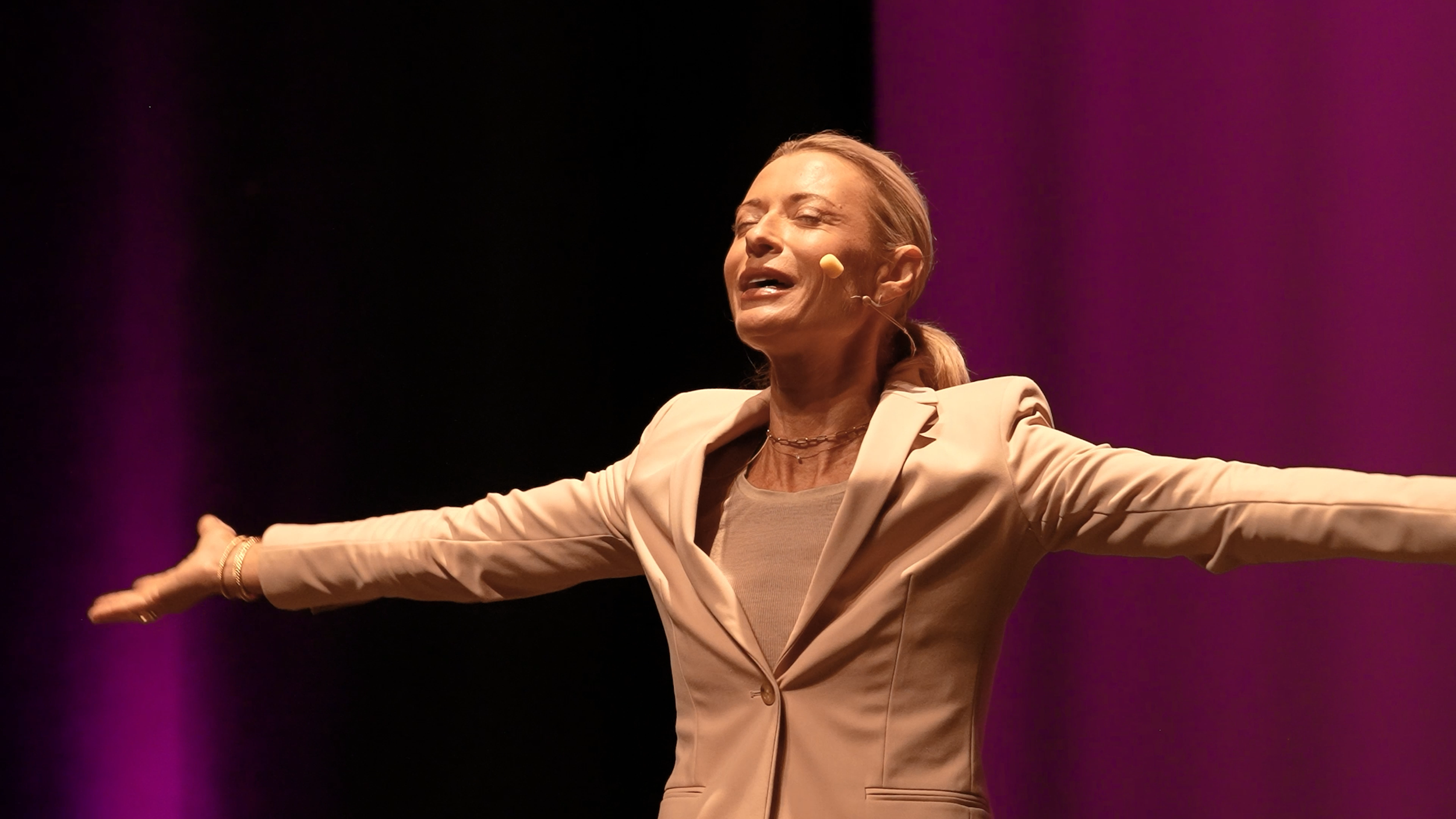 A woman wearing a beige blazer and top, with her arms outstretched and eyes closed, performing on stage with a purple background.