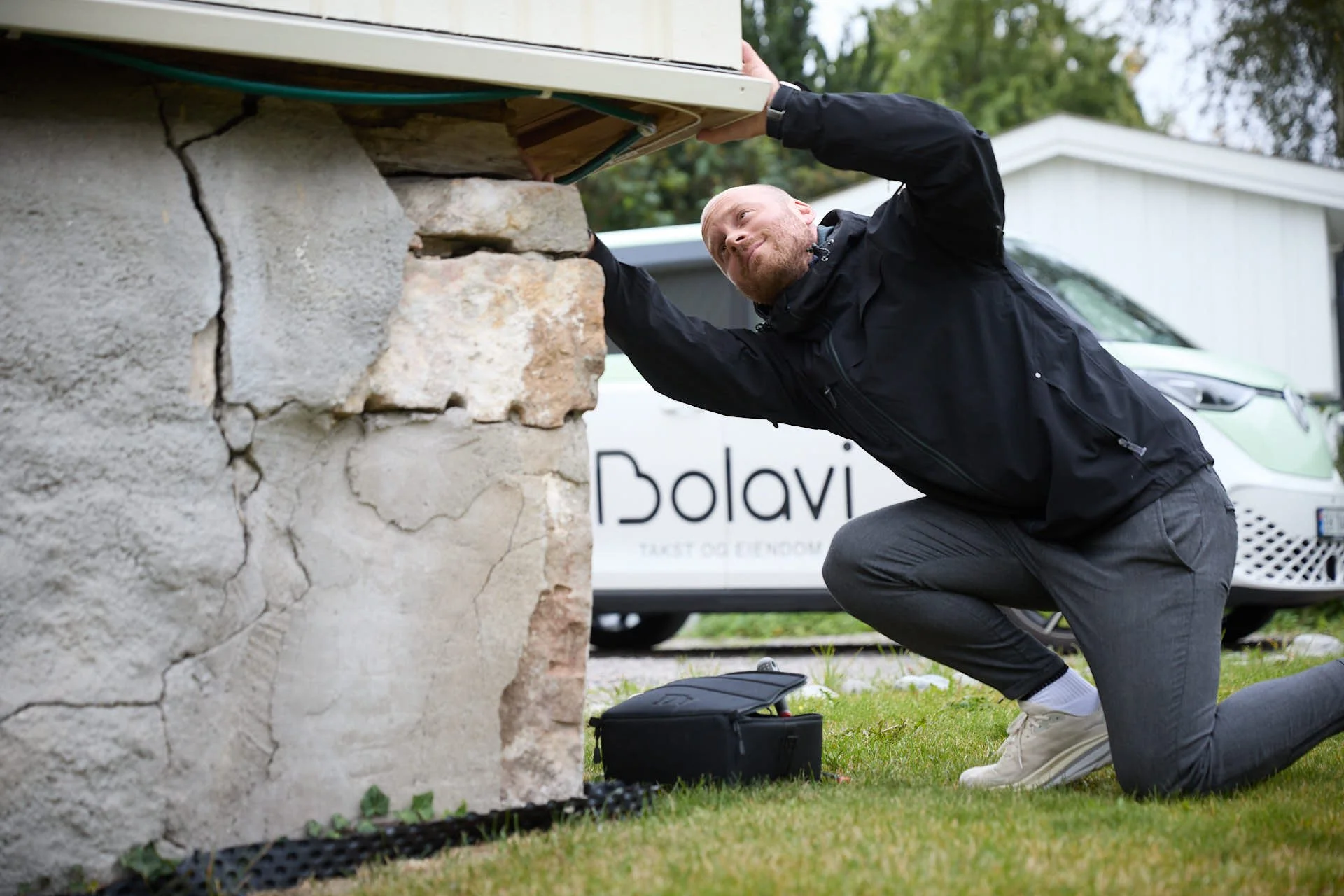 En mann i svart jakke og grå bukse som kneler på gresset og jobber med noe under et utbyggingsbjelke utendørs. I bakgrunnen en hvit bil med logoen 'Bolavi'.