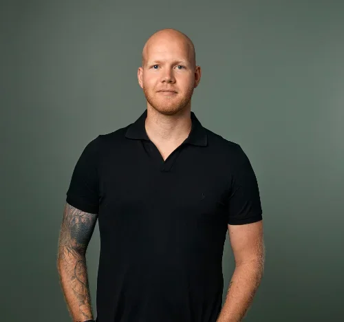 Mand med kort, lyst hår, tatovering på venstre arm, iført svart polo skjorte, uten smil, stående mot en grå bakgrunn.