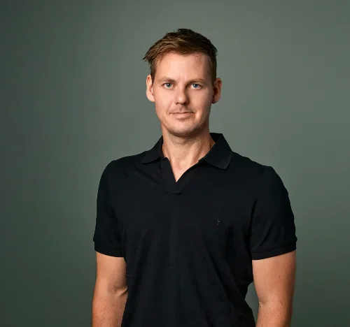 Portrett av en mann med kort, lysbrunt hår iført en svart polo-shirt, stående mot en grå bakgrunn.