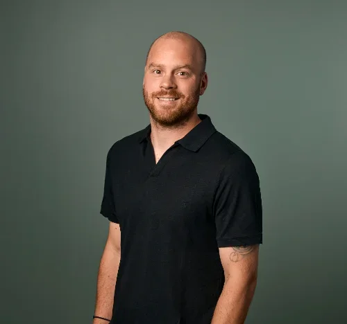 En mann med skjegg i svart polo-shirt smiler mot et grått bakgrunn.