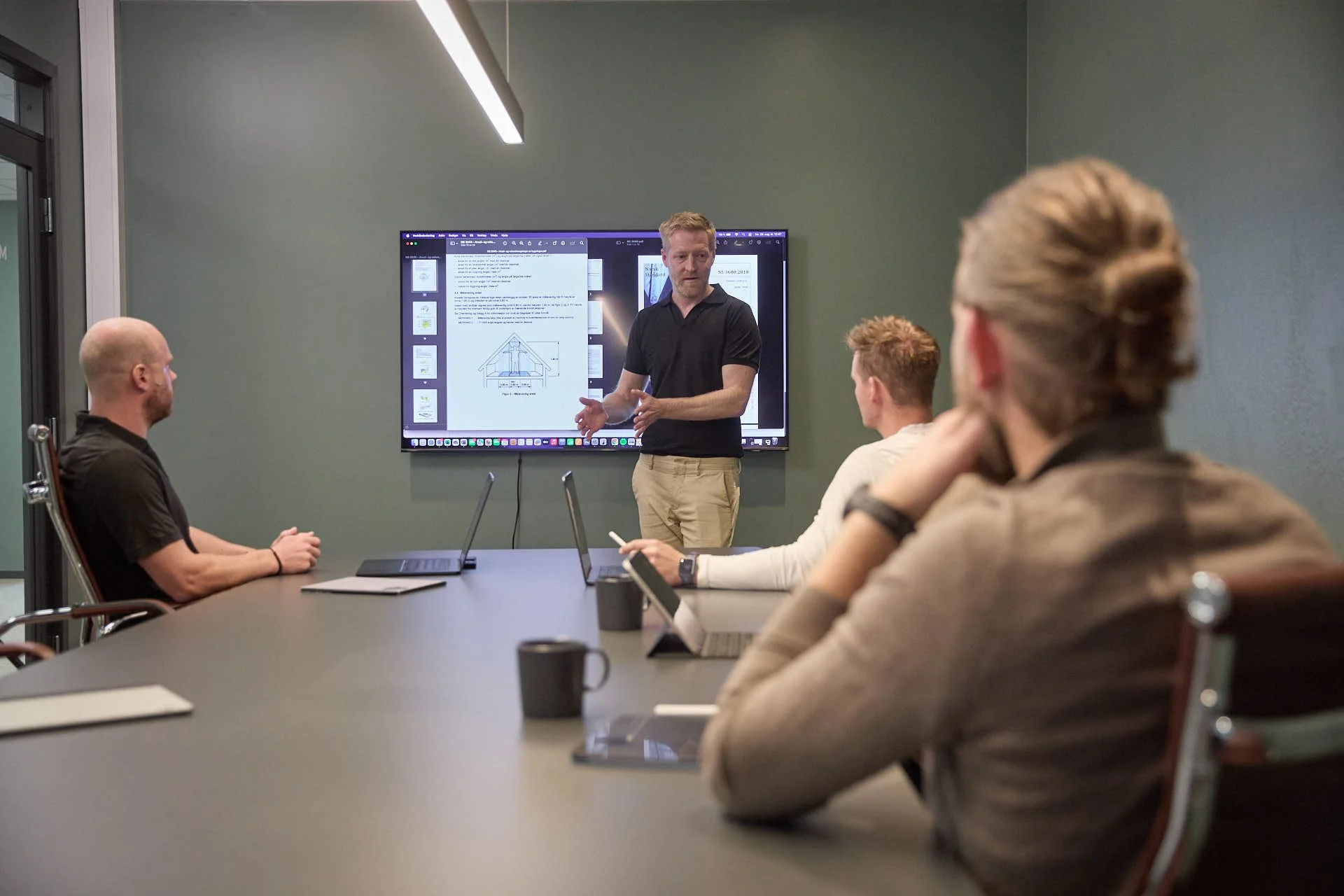 En mann holder en presentasjon foran fire personer i et møterom med en stor skjerm som viser en digital presentasjon av et arkitektonisk tegning.